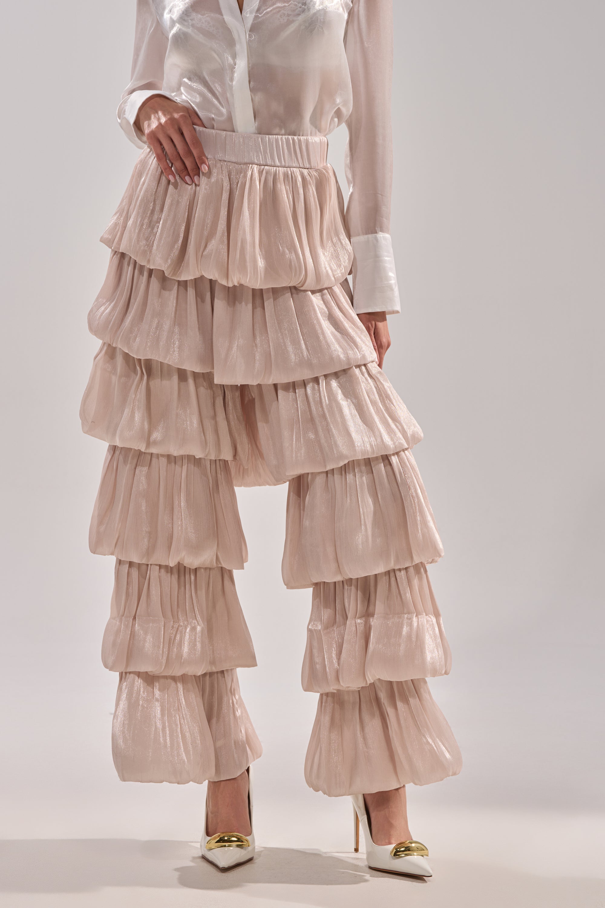 A woman models the STAR OF THE NIGHT FLOUNCY PANT IN IVORY—high-rise, tiered ruffle pants—paired with a sheer white blouse and white heels with gold accents; image cropped at shoulders to spotlight the outfit. 1