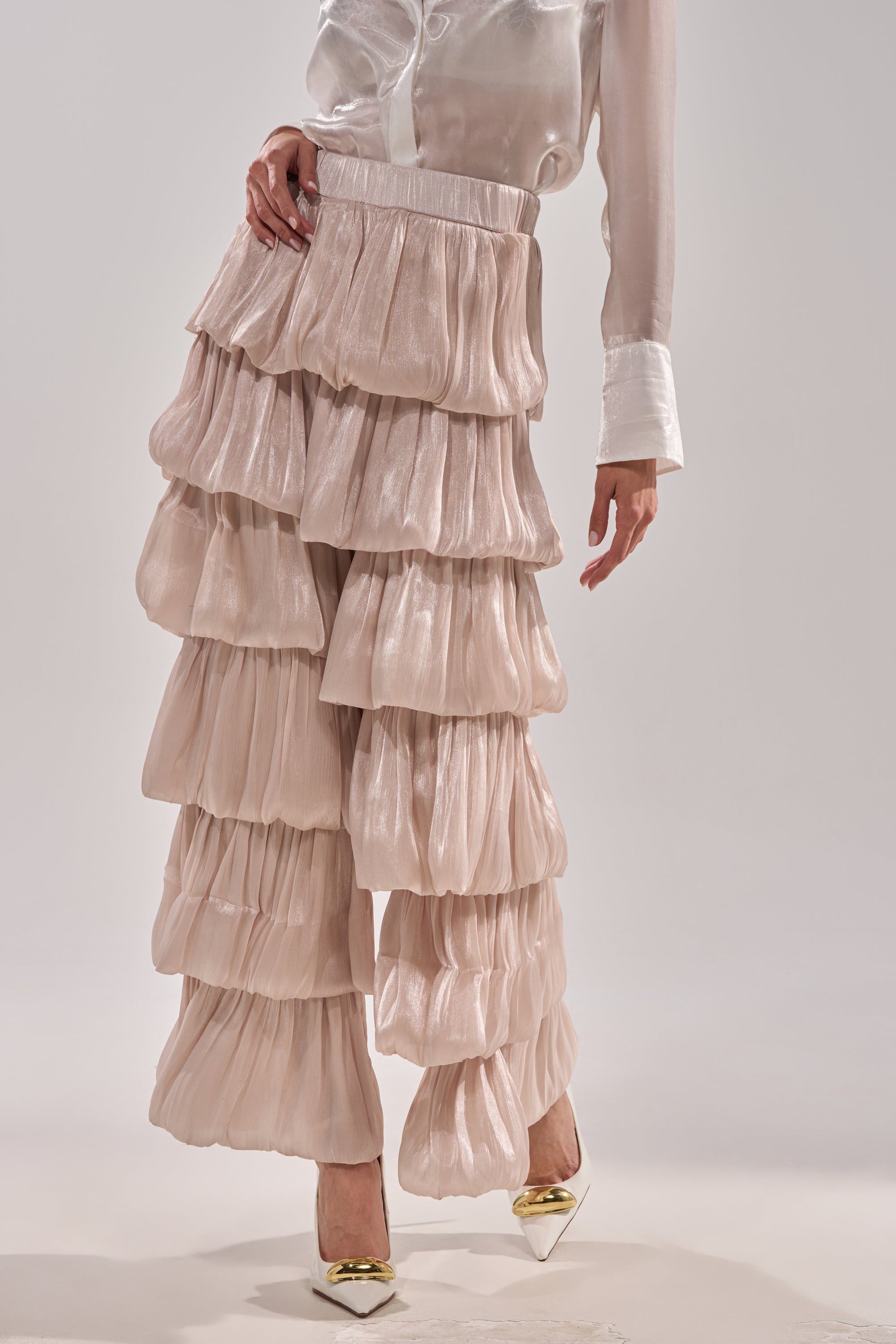 A person stands on a plain background wearing the STAR OF THE NIGHT FLOUNCY PANT IN IVORY and a sheer, long-sleeve blouse with white cuffs, styled with pointed white heels featuring metallic gold tips. 0