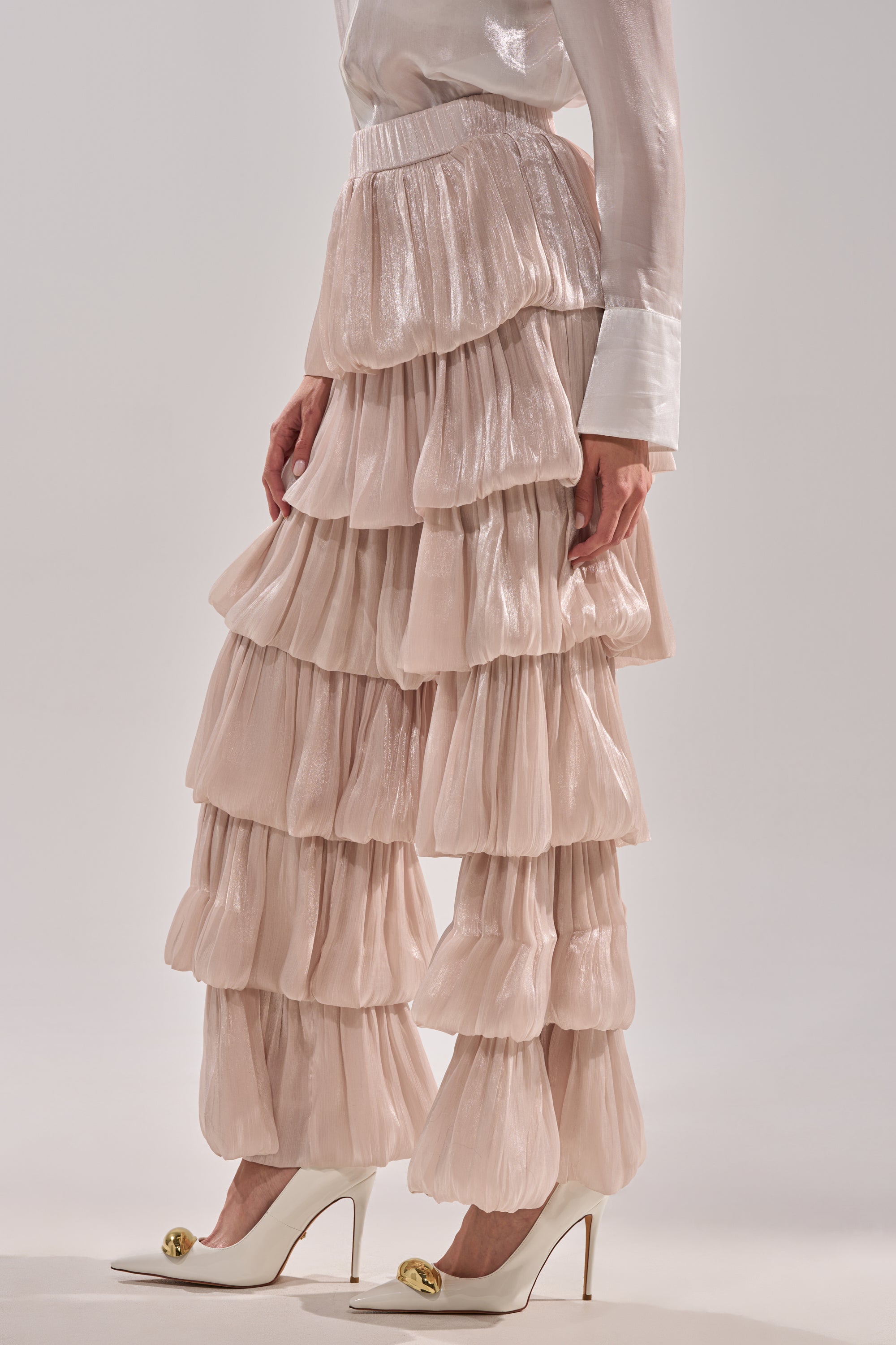 A person wearing a white, long-sleeved, shiny blouse and the STAR OF THE NIGHT FLOUNCY PANT IN IVORY, paired with white high-heeled shoes featuring gold embellishments. 2