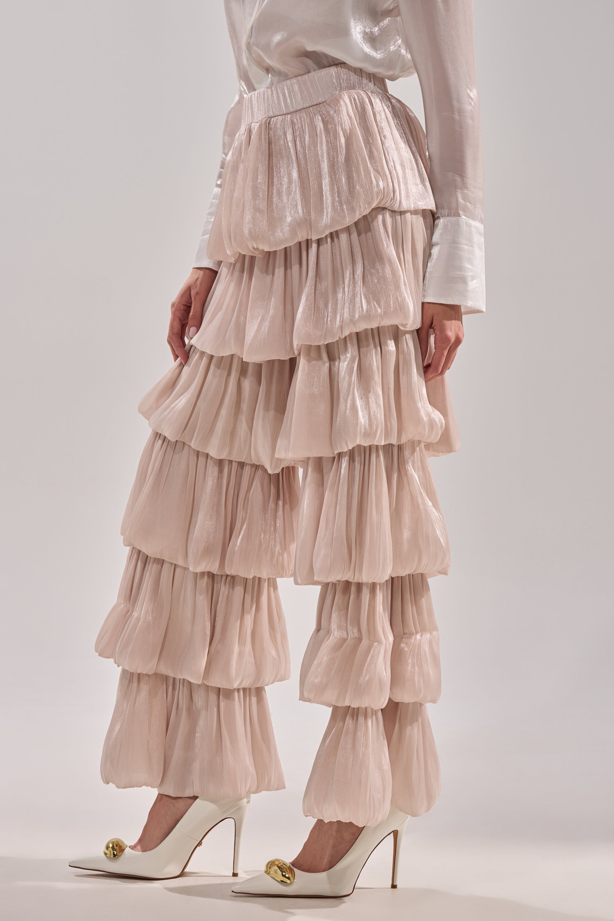 Wearing the STAR OF THE NIGHT FLOUNCY PANT IN IVORY with a matching long-sleeve blouse and white high heels adorned with gold toe embellishments, a person stands elegantly against a plain background. 3