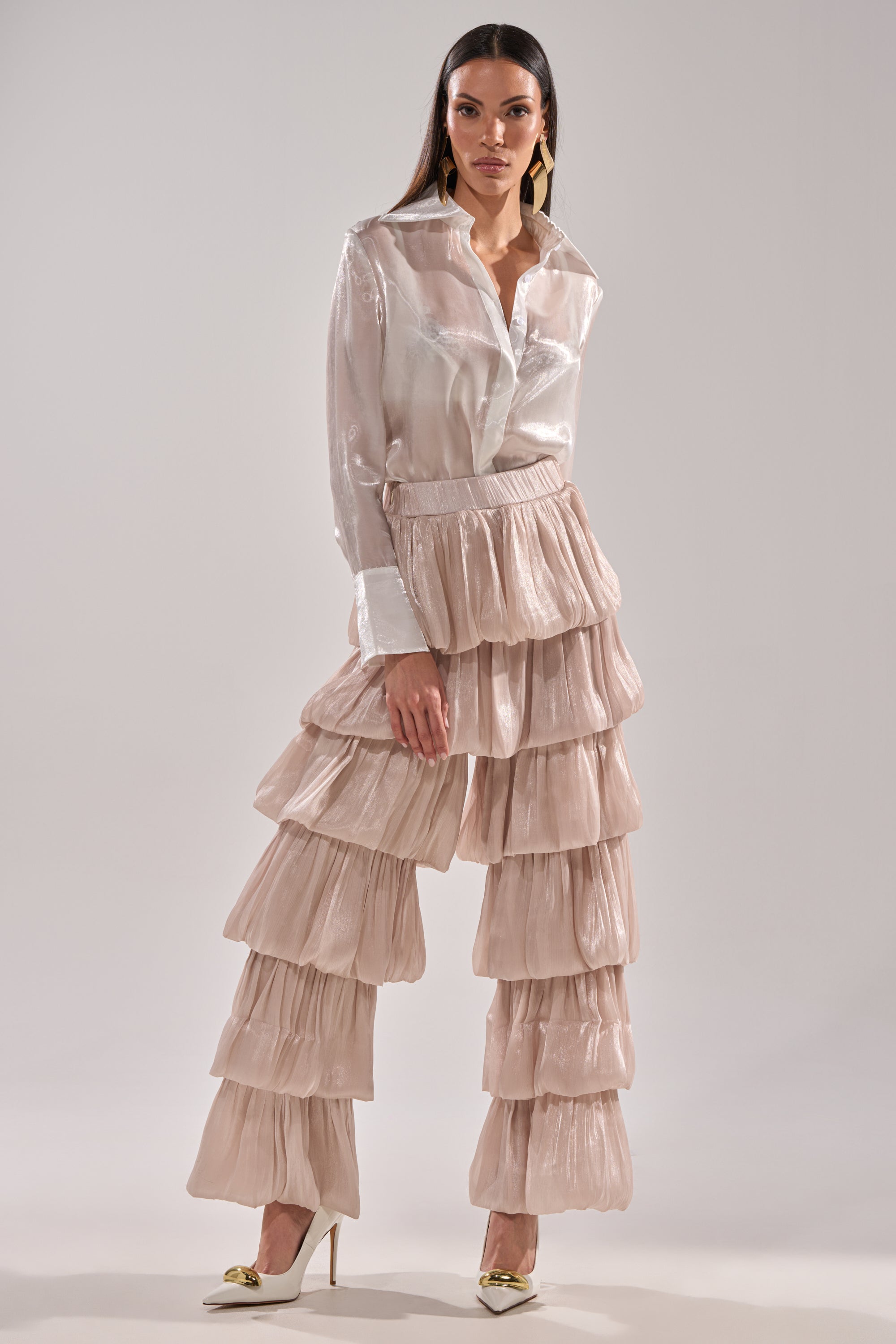 A woman poses in STAR OF THE NIGHT Flouncy Pant in Ivory, featuring a ruffled wide-leg silhouette, paired with a sheer white blouse and white heels against a plain light gray background. 6