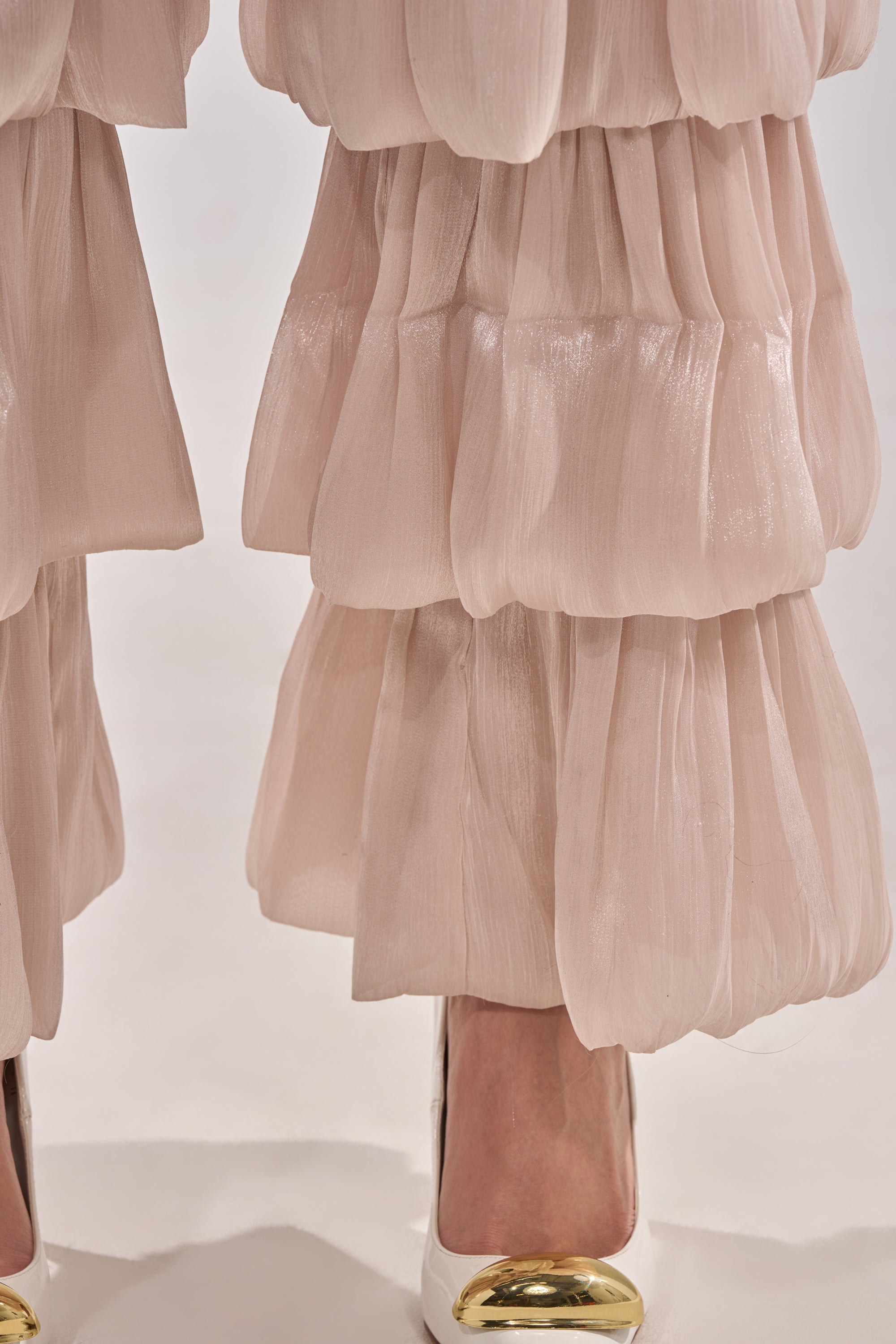 Close-up of a person wearing STAR OF THE NIGHT FLOUNCY PANT IN IVORY—layered, pleated wide-leg organza pants—paired with white high heels with gold metallic toe caps, standing on a light surface. 8