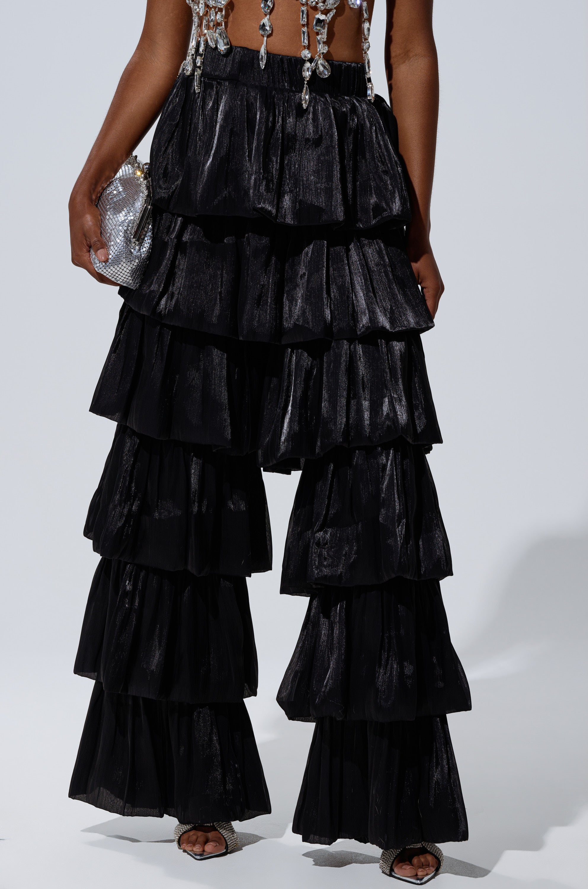 A person wears the STAR OF THE NIGHT FLOUNCY PANT IN BLACK—dramatic, tiered ruffle, wide-leg pants—paired with a beaded top and small silver handbag. Only their lower half is shown against a plain background. 0