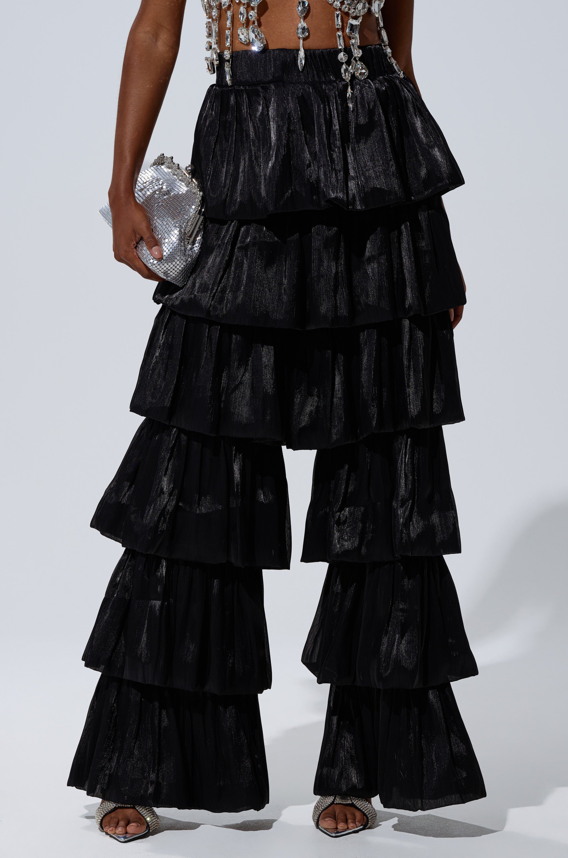 Wearing the STAR OF THE NIGHT FLOUNCY PANT IN BLACK with dramatic tiered ruffles and wide legs, a person holds a silver textured clutch and pairs the look with an embellished cropped top against a plain, well-lit background. 1