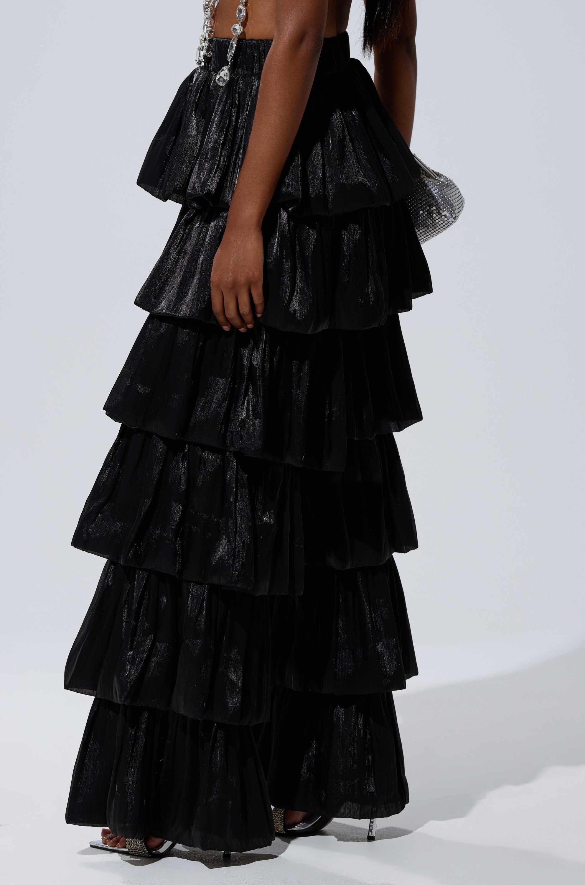 Wearing the STAR OF THE NIGHT FLOUNCY PANT IN BLACK, a person showcases its wide-leg silhouette against a plain background. Only their lower half is visible, paired with open-toe heels and a small textured bag. 2