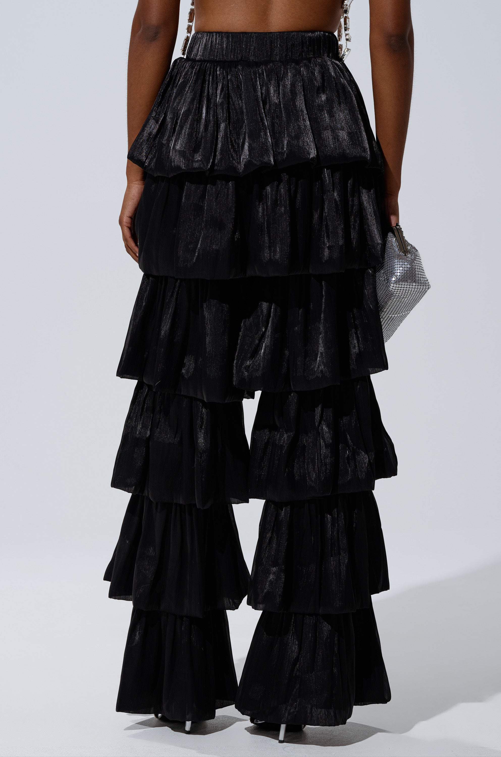 Photographed from the back against a plain light background, a person wears the STAR OF THE NIGHT FLOUNCY PANT IN BLACK—tiered ruffle, wide-leg pants—while holding a small silver clutch. 3
