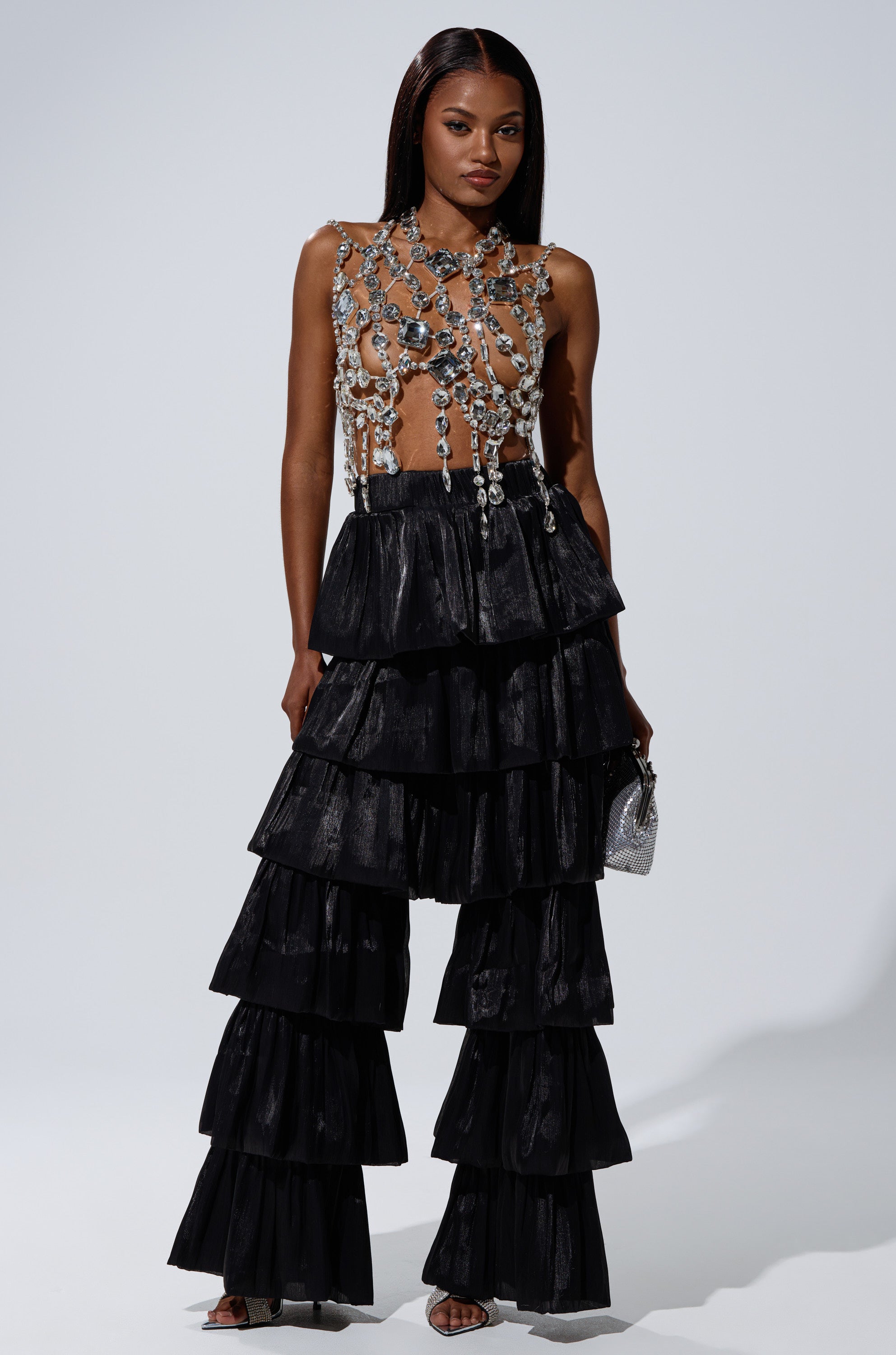 A woman wears the STAR OF THE NIGHT FLOUNCY PANT IN BLACK—a dramatic, wide-leg, tiered ruffle pant—paired with a sleeveless top accented by silver chains and crystals. She completes the look with silver heels and a small silver clutch against a plain light backdrop. 4