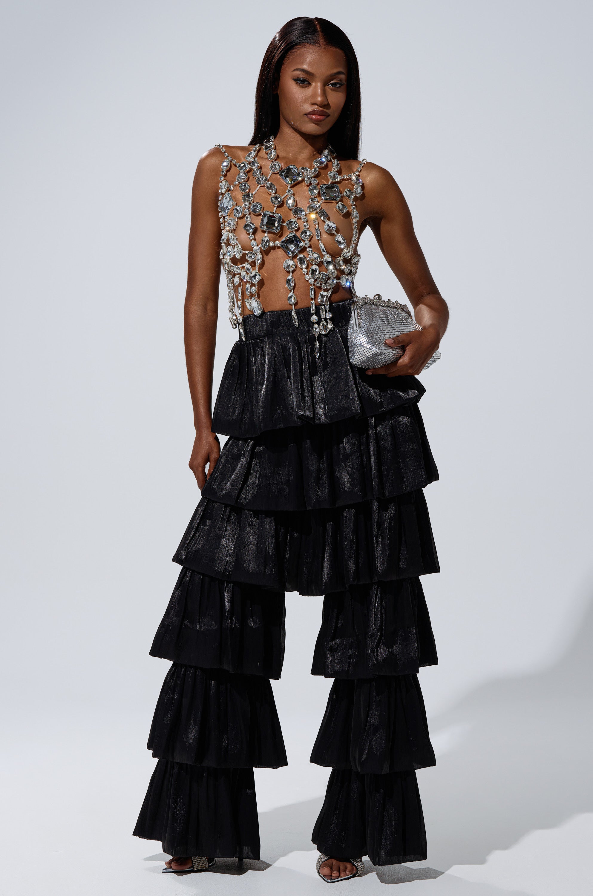 A woman poses confidently against a light background in the STAR OF THE NIGHT FLOUNCY PANT IN BLACK, featuring tiered ruffles. She pairs it with a sheer bejeweled top, holds a silver clutch, and wears her hair straight and sleek. 5