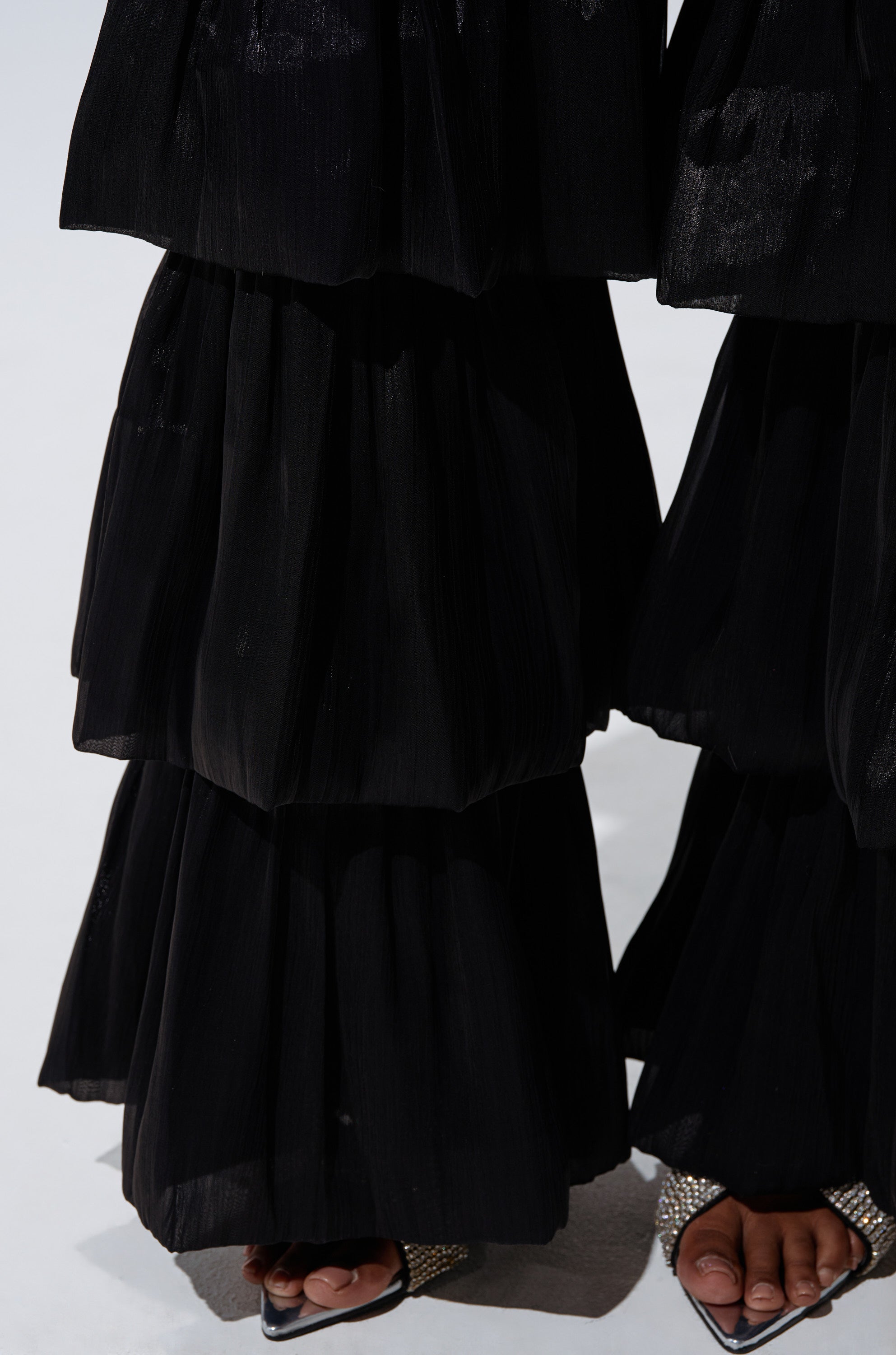 A close-up shows only the lower legs and feet of a person wearing the STAR OF THE NIGHT FLOUNCY PANT IN BLACK and open-toe heels with rhinestone embellishments, standing on a white surface. 6