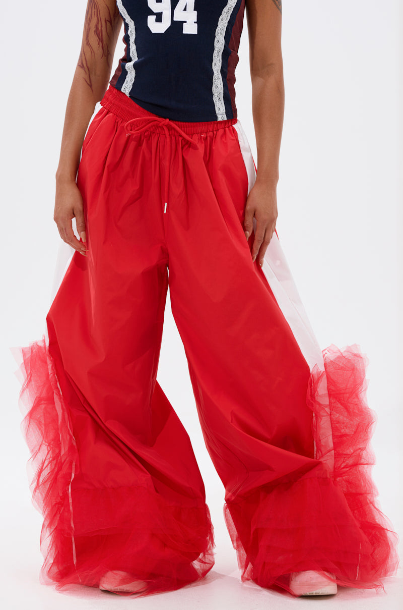Cropped photo shows a person in FEELING LIKE A BOSS TRACK PANT WITH TULLE TRIM IN RED featuring wide legs and ruffled tulle hems, paired with a navy sleeveless top with white lace trim and the number 94. 0