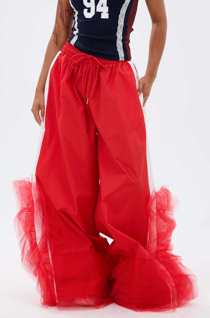 A person wears the FEELING LIKE A BOSS TRACK PANT WITH TULLE TRIM IN RED, featuring wide legs and tulle ruffles, styled with a sleeveless navy top with a white number 94. Photo cropped from shoulders to ankles. 1