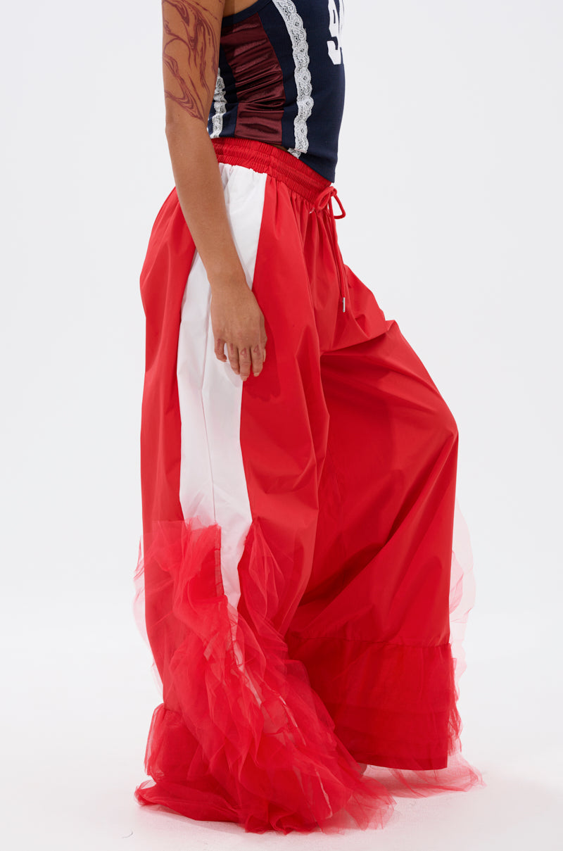 Wearing FEELING LIKE A BOSS TRACK PANT WITH TULLE TRIM IN RED, featuring white stripes and tulle detail, a person with a tattooed arm stands against a white background, paired with a navy top. 2
