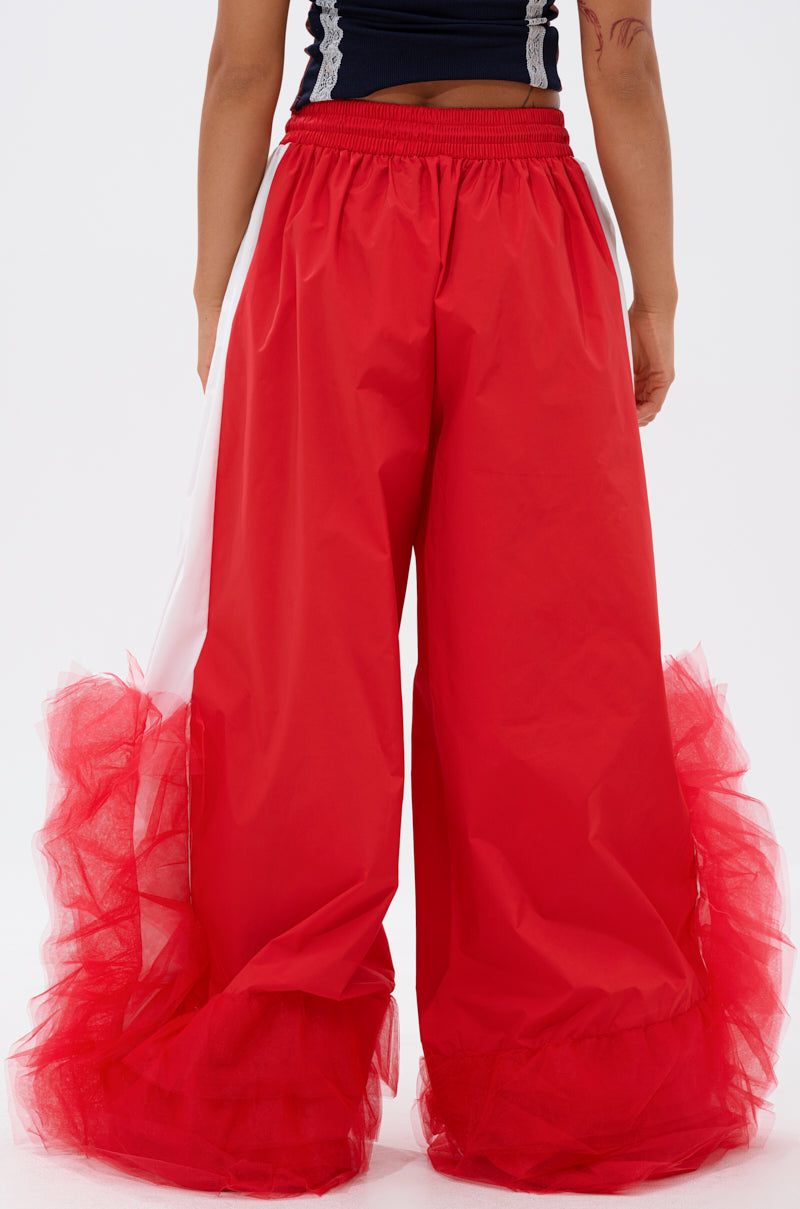 A model showcases the FEELING LIKE A BOSS TRACK PANT WITH TULLE TRIM IN RED, featuring wide legs and tulle ruffle accents at the hem, shown from the waist down against a plain white background. 3