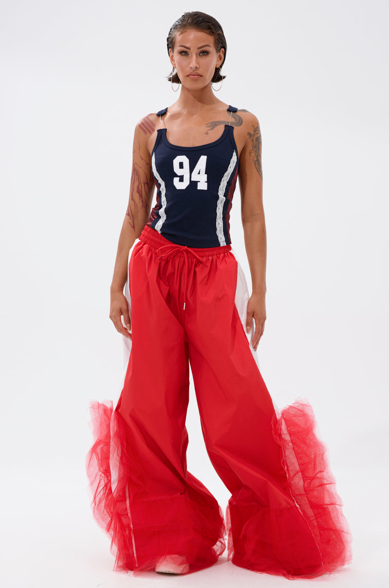 A person stands against a plain background wearing FEELING LIKE A BOSS TRACK PANT WITH TULLE TRIM IN RED, featuring bright red wide-leg legs and sheer tulle trim along the sides and bottoms, paired with a navy tank top displaying "94. 4