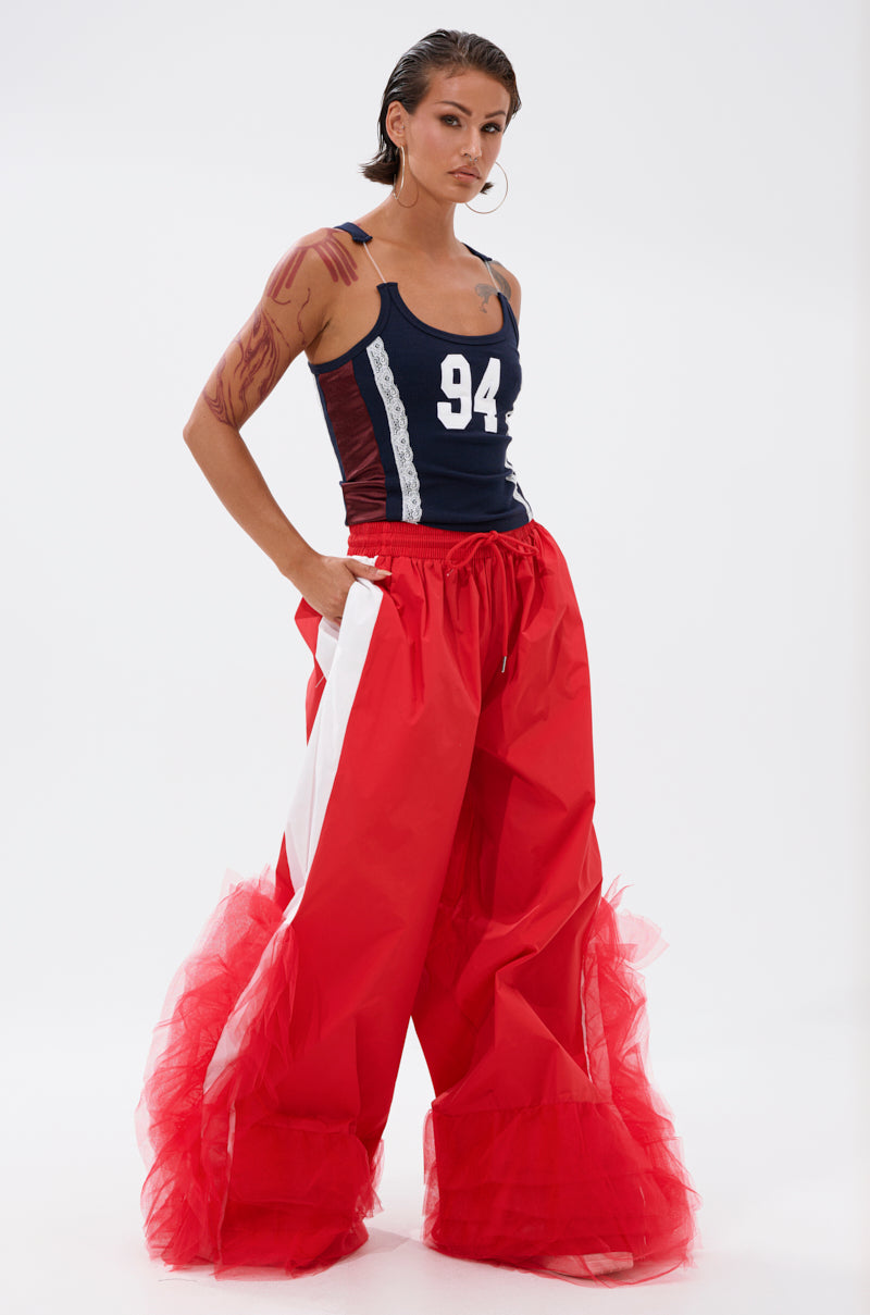 A person with slicked-back hair poses confidently in FEELING LIKE A BOSS TRACK PANT WITH TULLE TRIM IN RED—bright red wide-leg track pants with bold white stripes and striking tulle trim—paired with a navy tank top numbered 94. 5