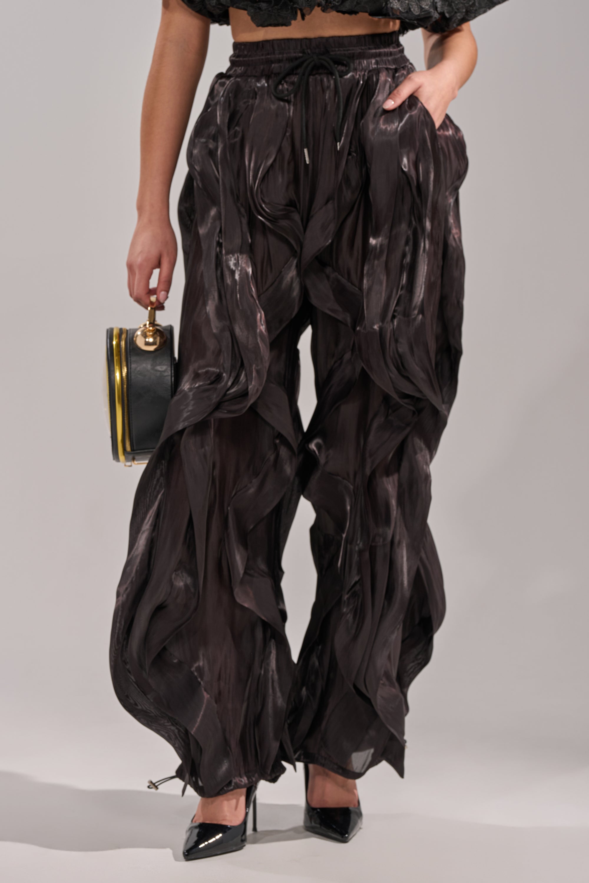 A person in avant-garde street style wears the COOLEST ON THE BLOCK JOGGER—loose, flowy, dark brown abstract pants—paired with a black cropped top, black pointed heels, and a small black handbag with a gold clasp against a plain background. 0