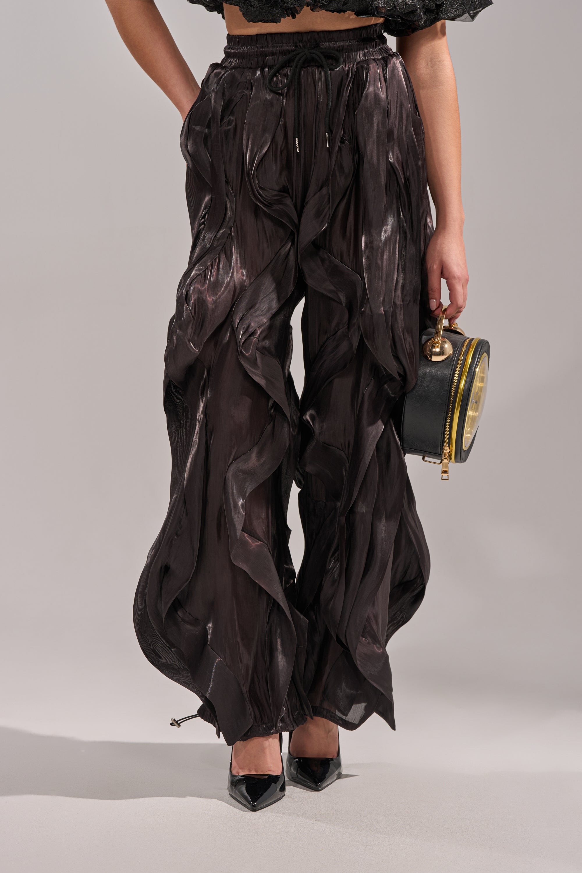 A person sports the COOLEST ON THE BLOCK JOGGER—shiny, dark, avant-garde wide-leg pants—with a cropped black top, black pointed heels, and a small black round handbag with a gold handle against a plain background. 1