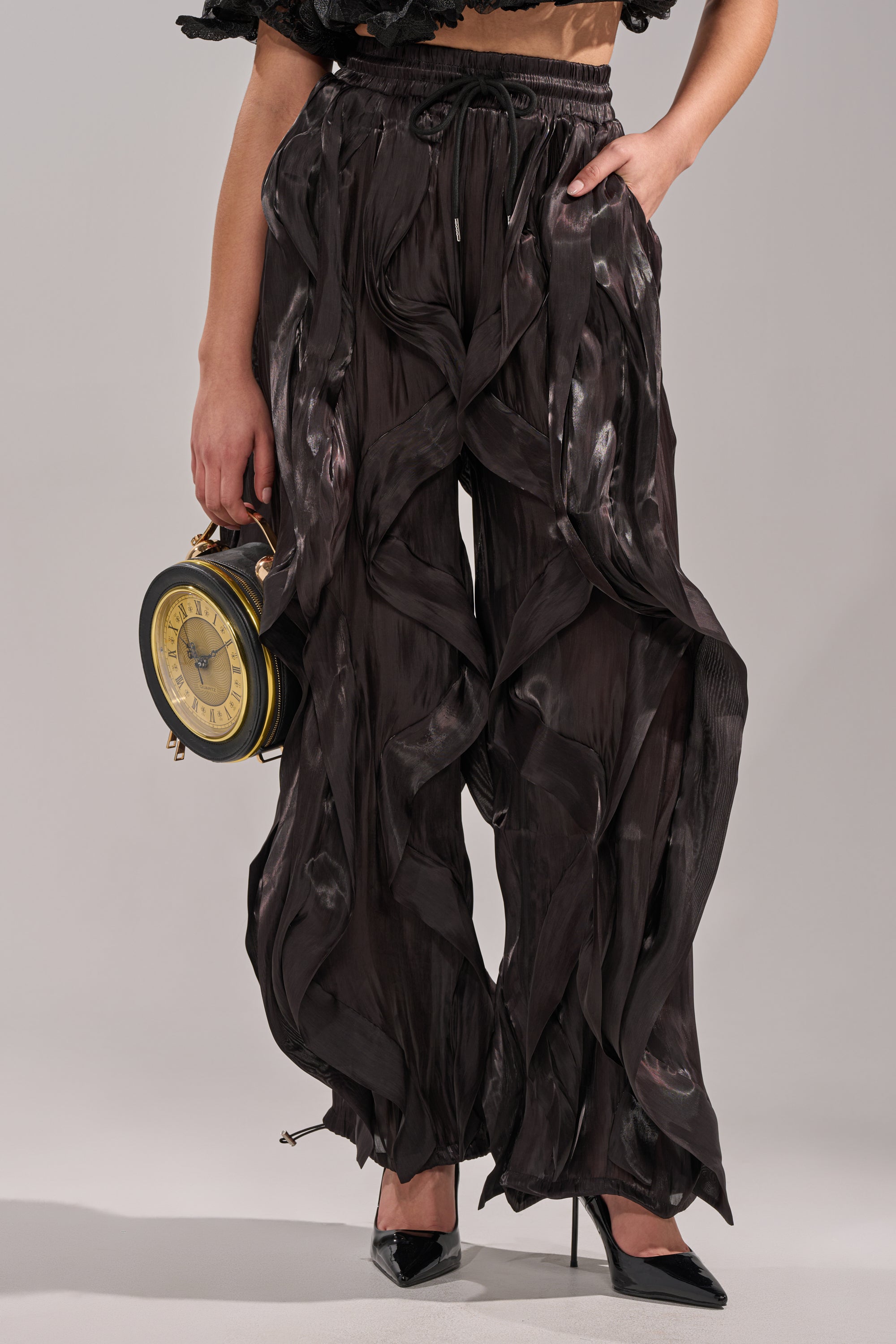 Wearing the COOLEST ON THE BLOCK JOGGER and black heels, a person holds a vintage clock-shaped handbag; only their lower half is visible against a plain background, showcasing avant-garde street style. 2