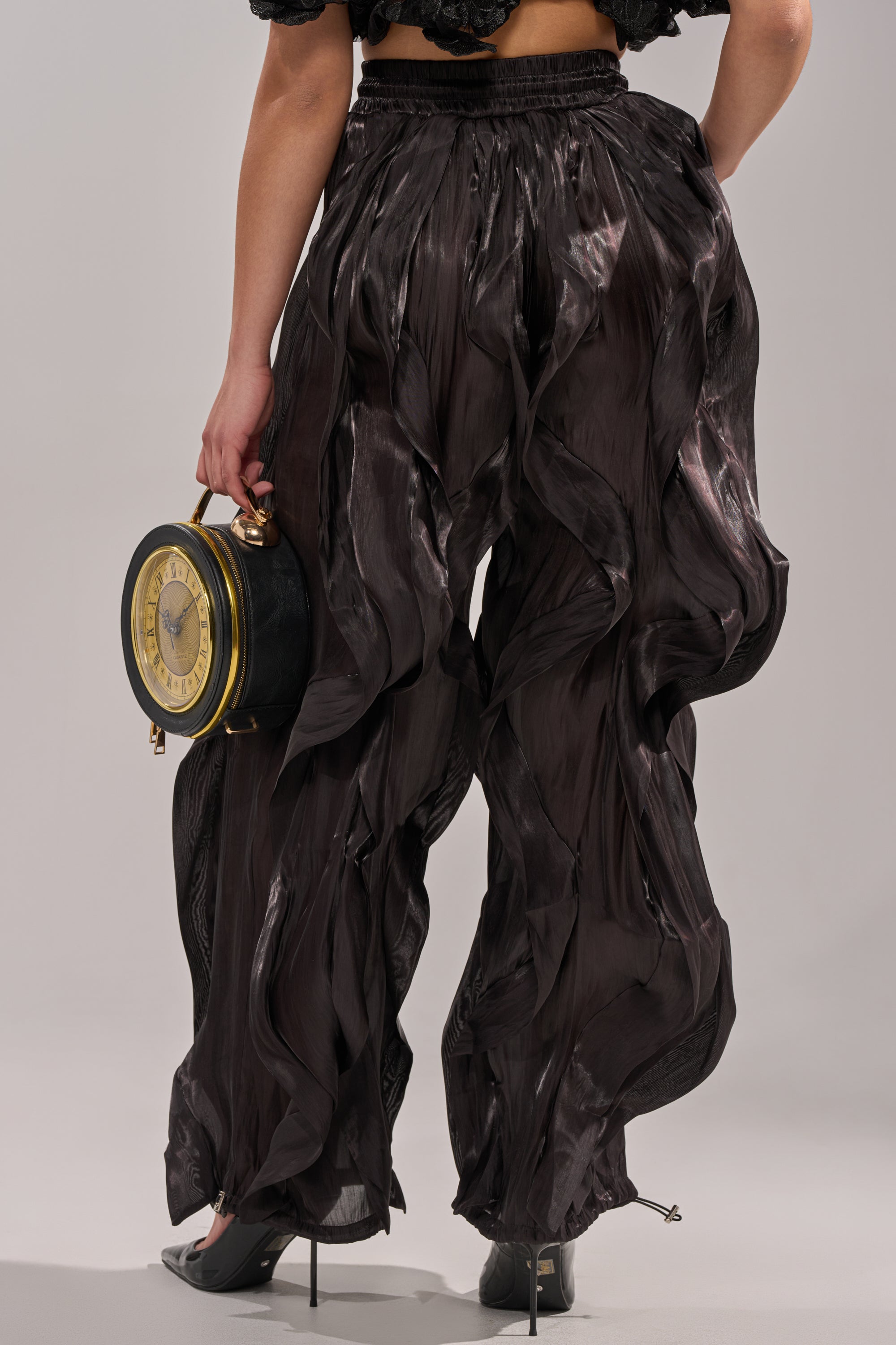 A person is seen from behind in COOLEST ON THE BLOCK JOGGER black sheer pants and heels, holding a clock-shaped round bag, with a plain light background. 4