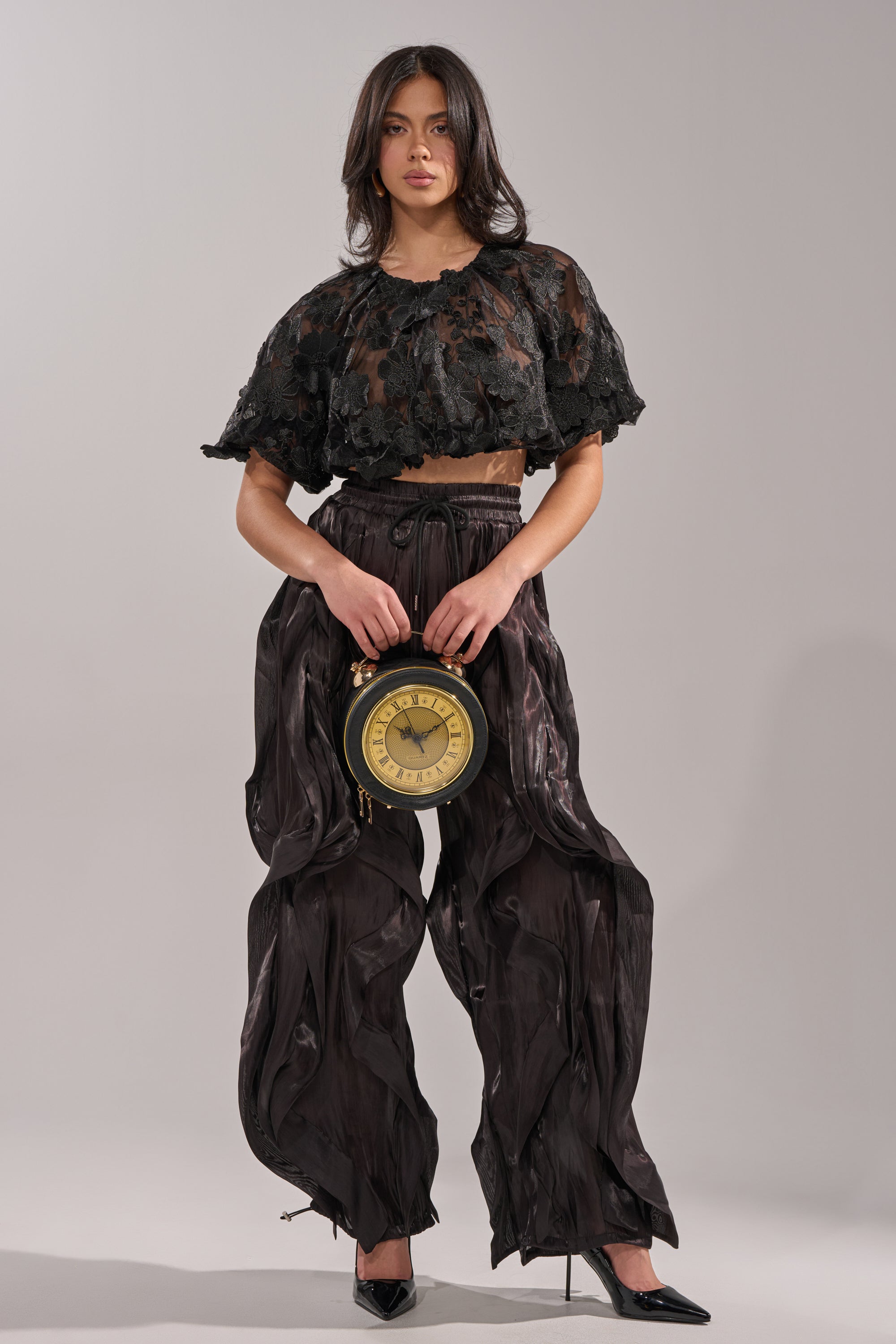 A woman in a sheer, ruffled black crop top and avant-garde street style COOLEST ON THE BLOCK JOGGER pants holds a vintage clock. She wears black heels and stands against a plain light gray background. 5