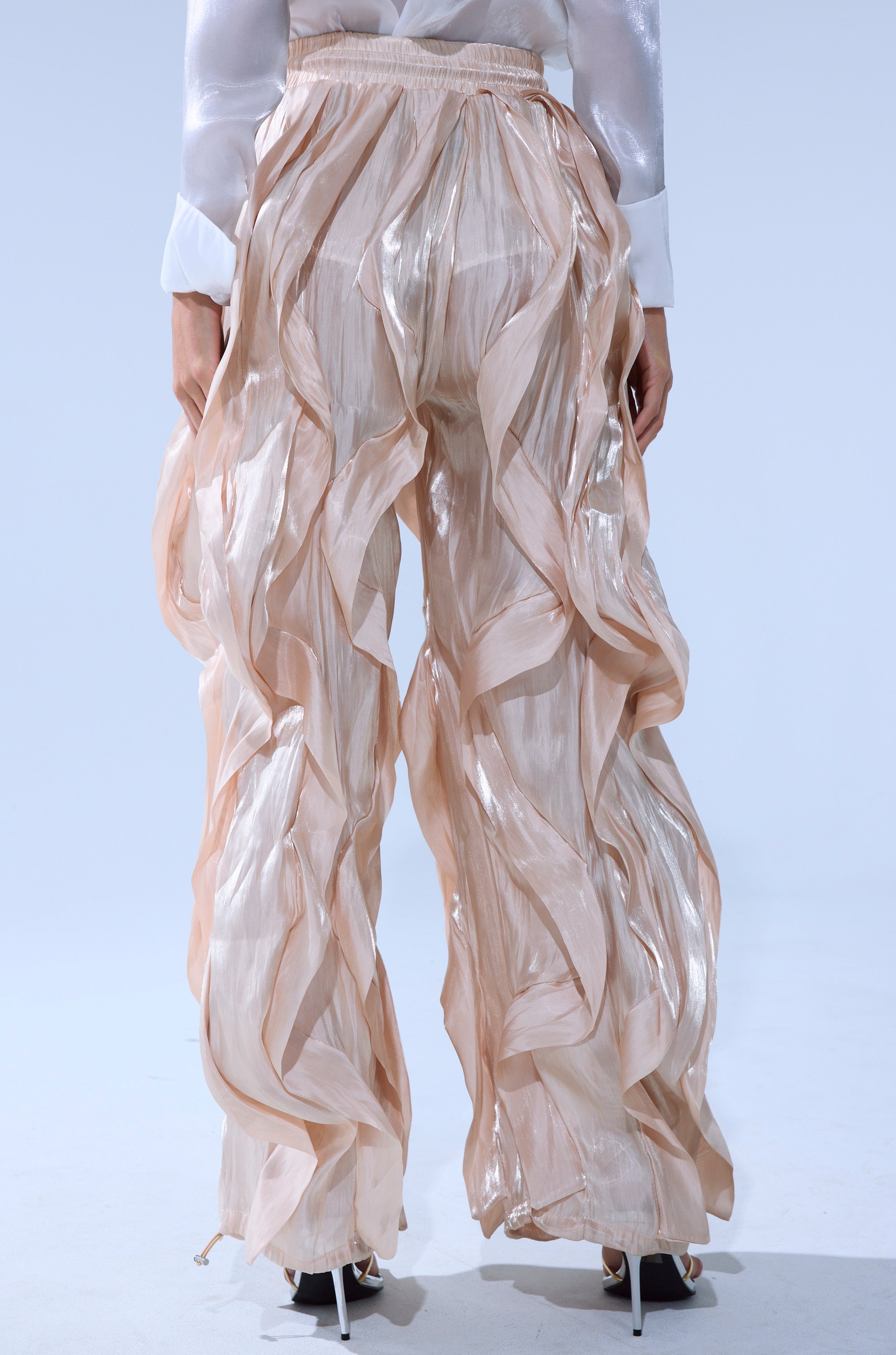 A person is shown from the back wearing the COOLEST ON THE BLOCK SATIN JOGGER—flowing sheer beige pants with an elastic waistband and ruffled layers—paired with a white blouse and heeled sandals, set against a plain light background.