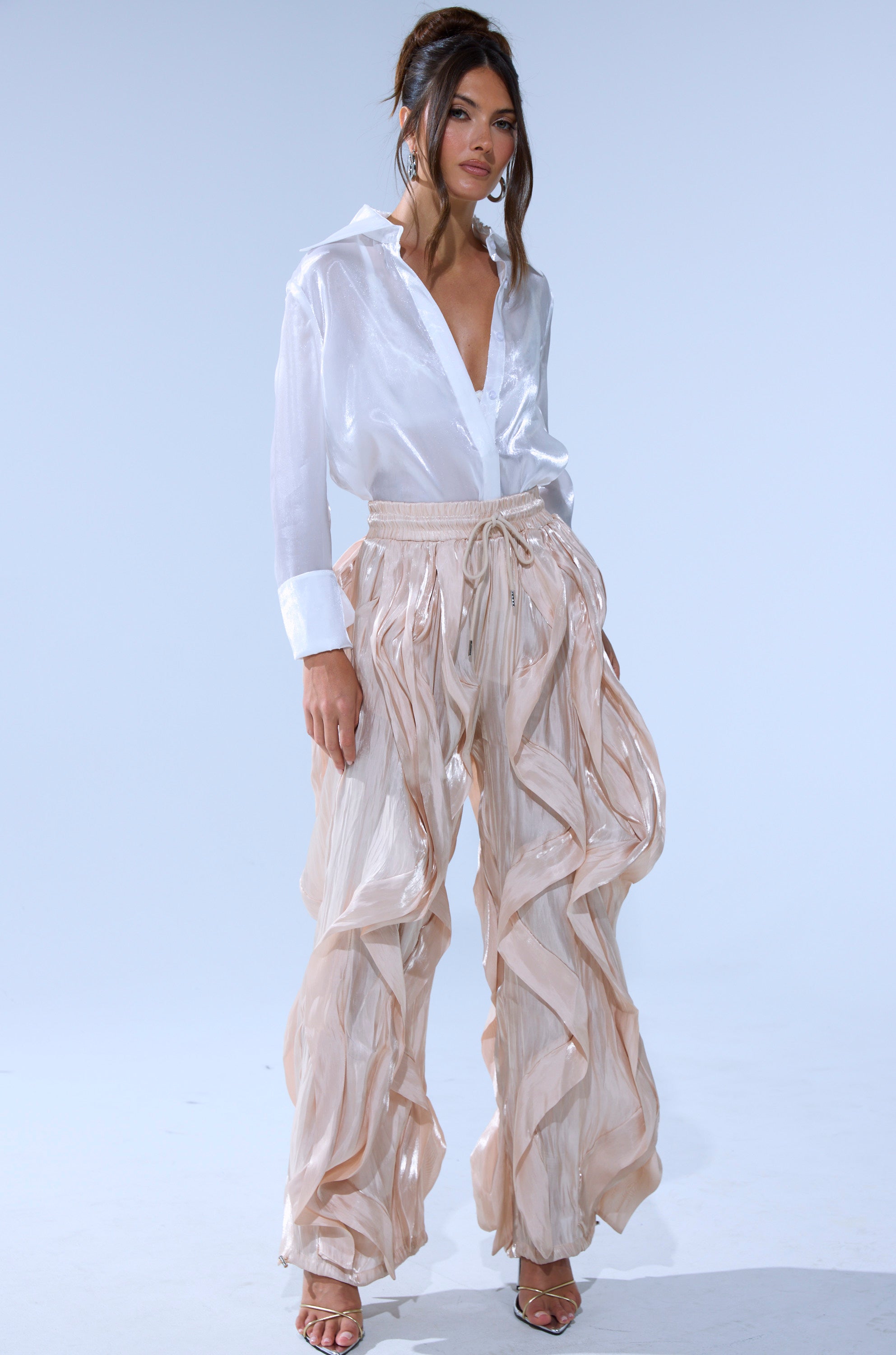 A woman stands against a plain background, wearing a shiny white button-down shirt and the COOLEST ON THE BLOCK SATIN JOGGER in beige. She has her hair up and pairs the look with strappy heels.