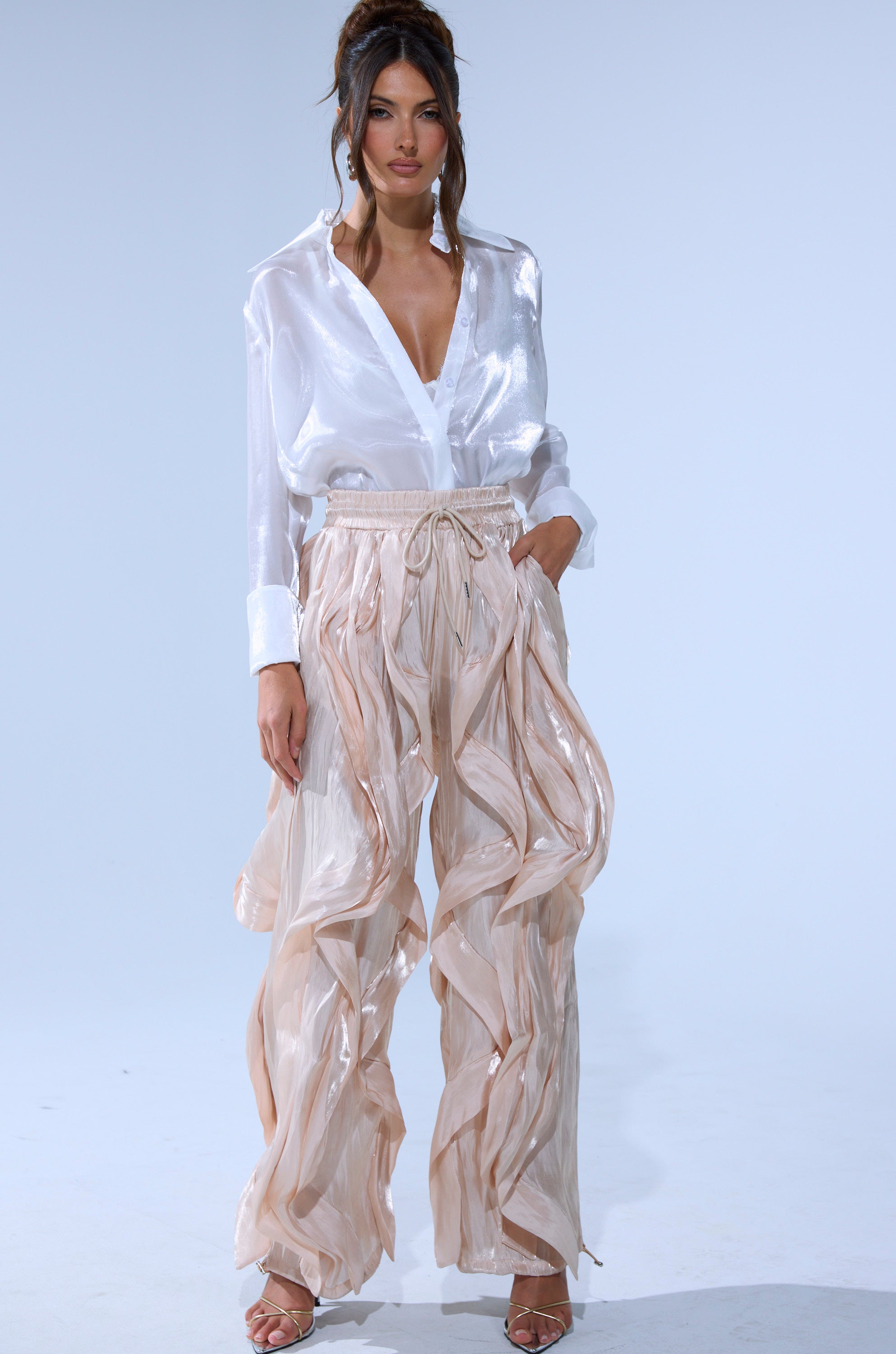 A woman stands against a plain light background wearing the COOLEST ON THE BLOCK SATIN JOGGER, styled with a shiny white blouse, hands in pockets, showcasing the jogger’s flowing wide-leg fit and ruffle details.