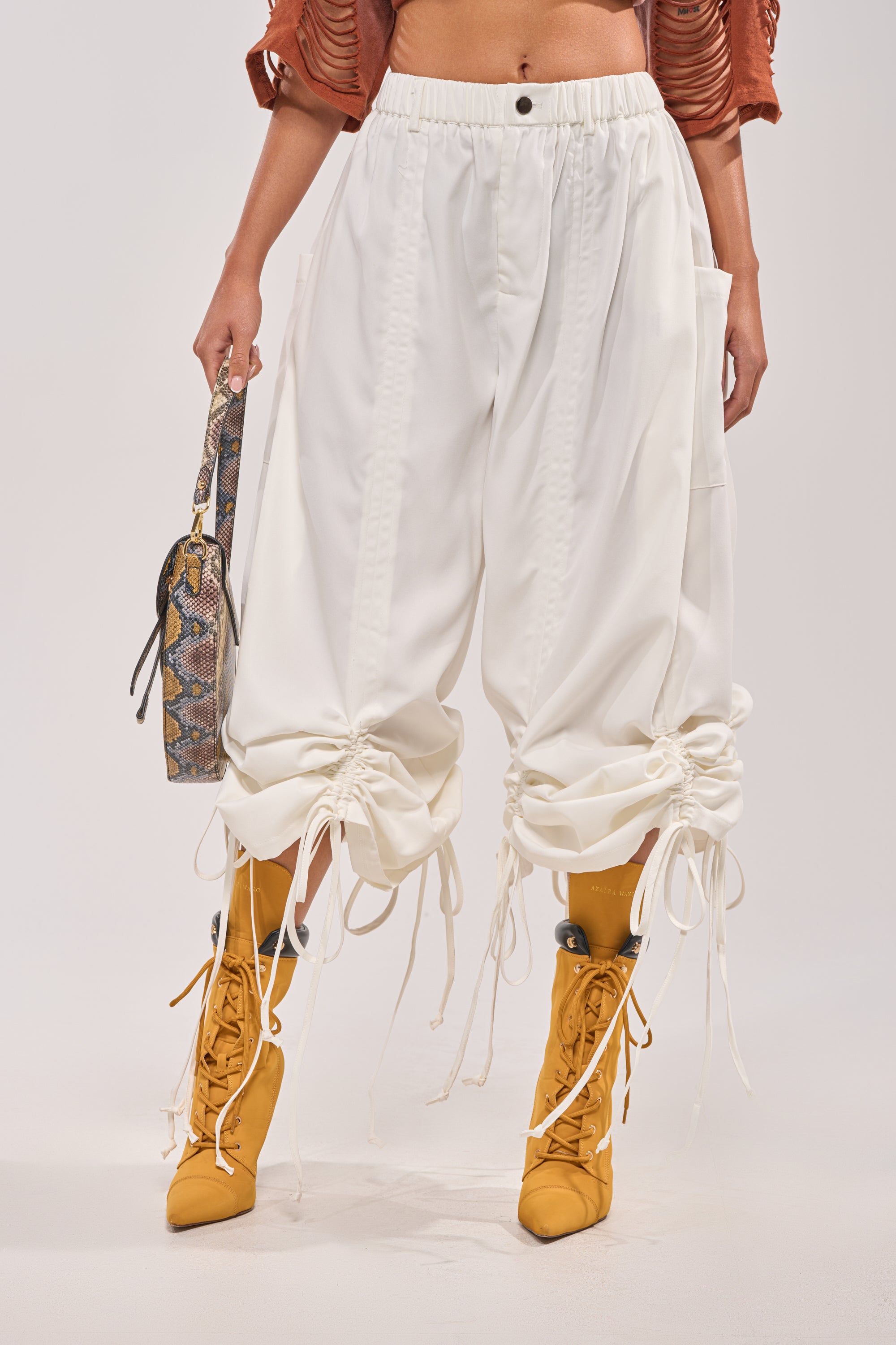 A person showcases street style in DRAMA RUCHED JOGGER IN WHITE, paired with mustard yellow lace-up boots, a cropped rust top, and a snakeskin-print handbag against a plain background. 0