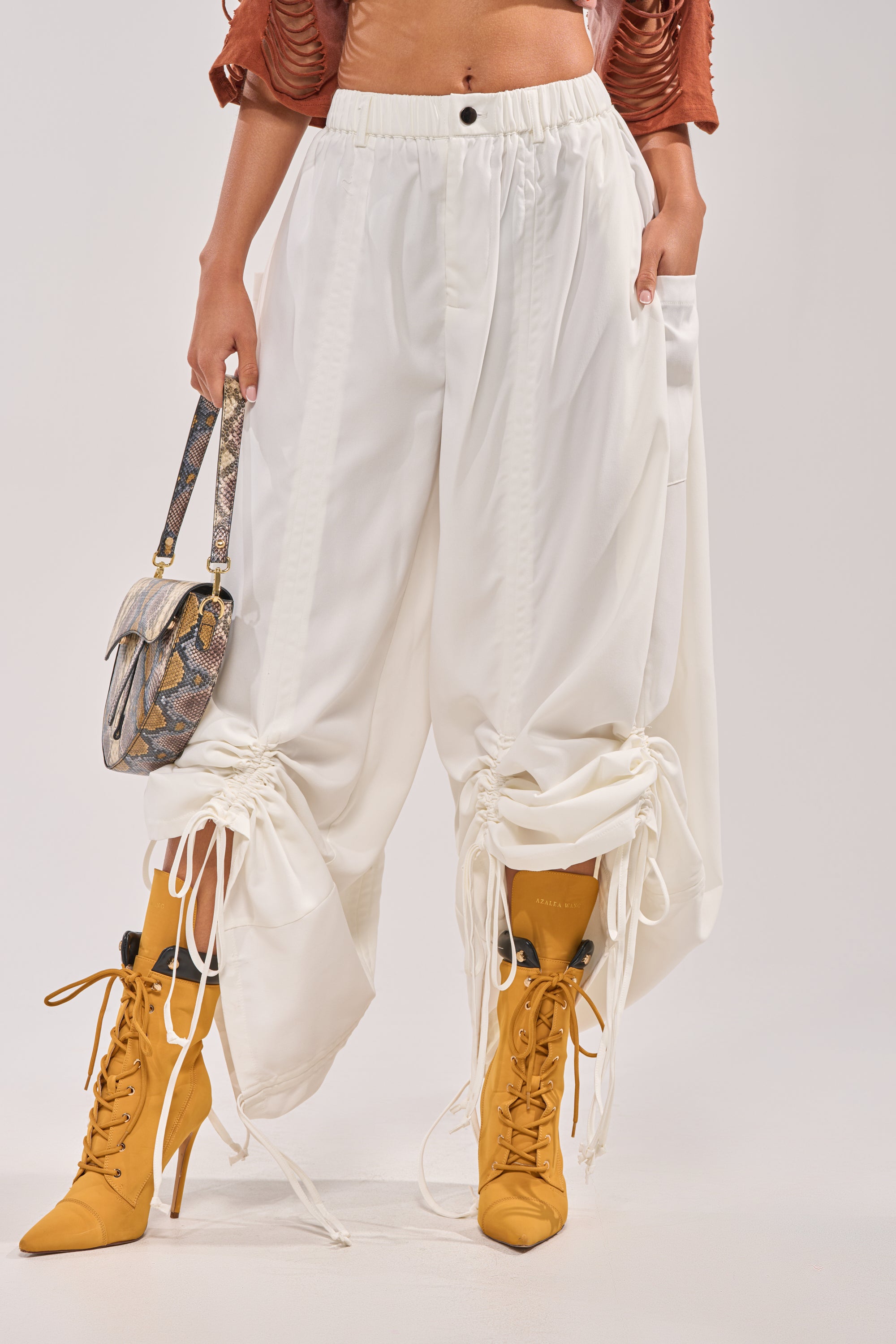 A person wears the DRAMA RUCHED JOGGER IN WHITE styled street-style with mustard yellow lace-up boots, a cropped rust top, and a small snakeskin-patterned handbag against a plain, light-colored background. 1