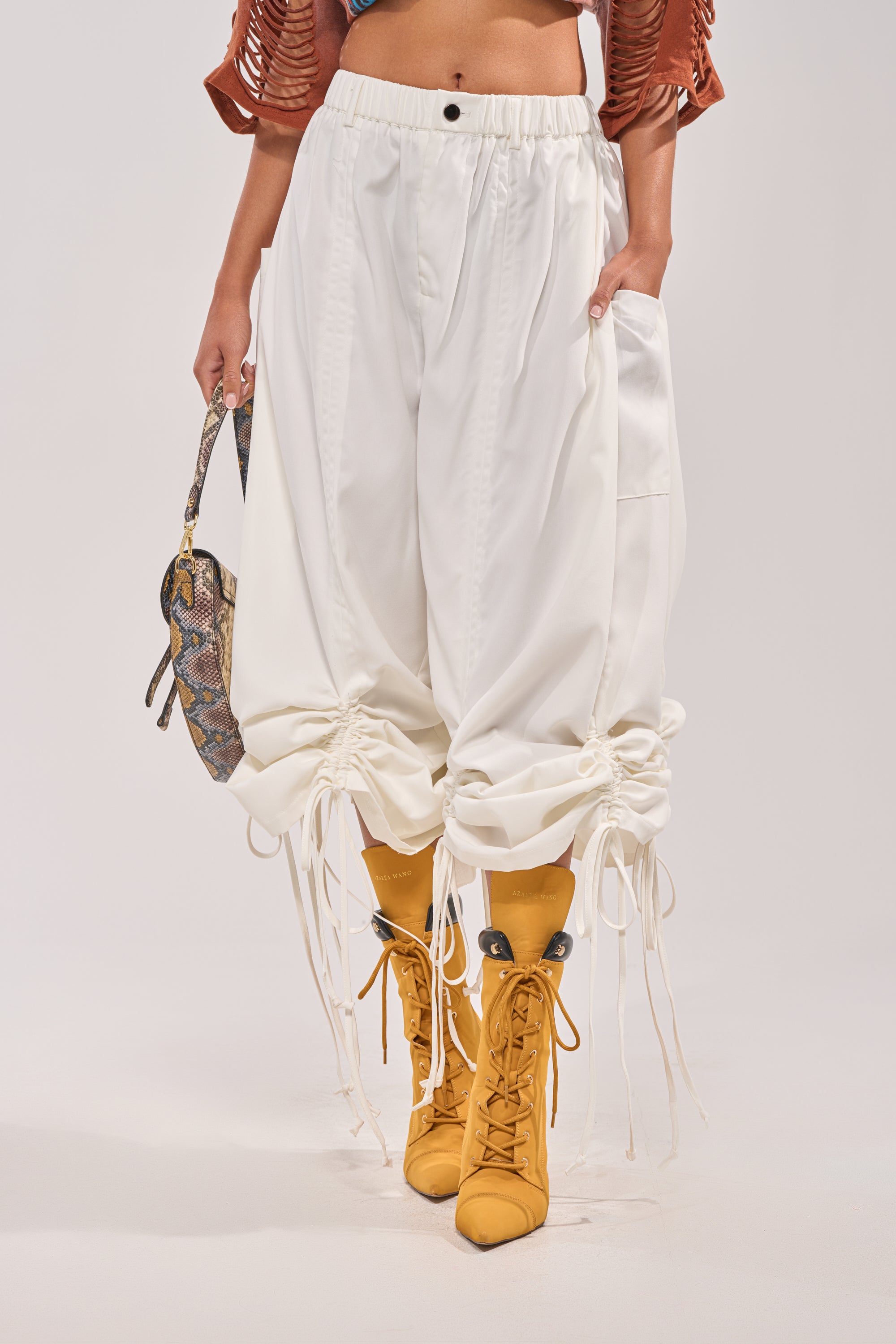 Wearing the DRAMA RUCHED JOGGER IN WHITE—high-rise with a button and elastic waistband—paired with yellow lace-up boots and a textured multicolored handbag. 2