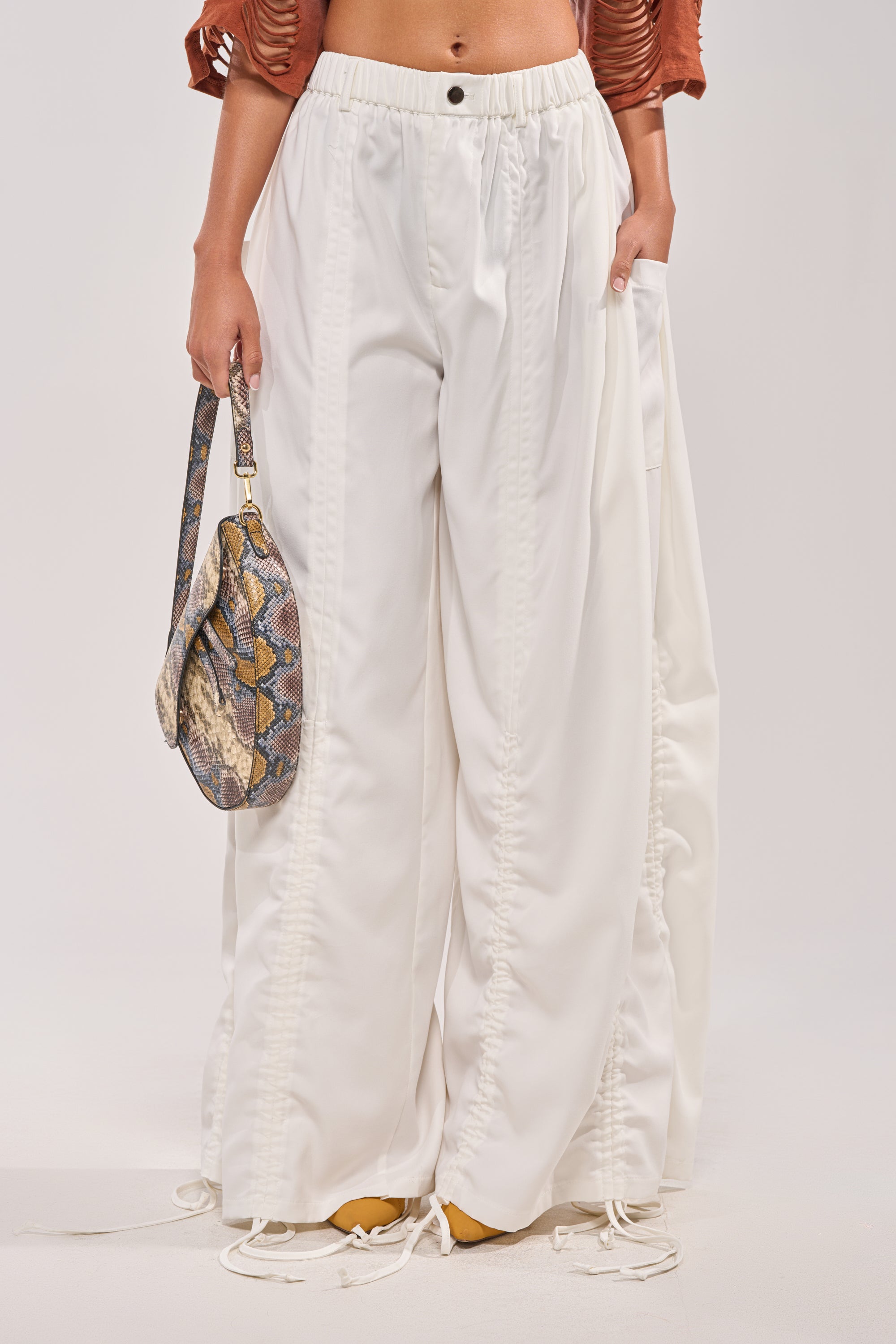 A person wears the DRAMA RUCHED JOGGER IN WHITE—loose, high-rise jogger pants with ruched details—while holding a snakeskin-print handbag. Only their lower torso and legs appear against a plain light background. 3