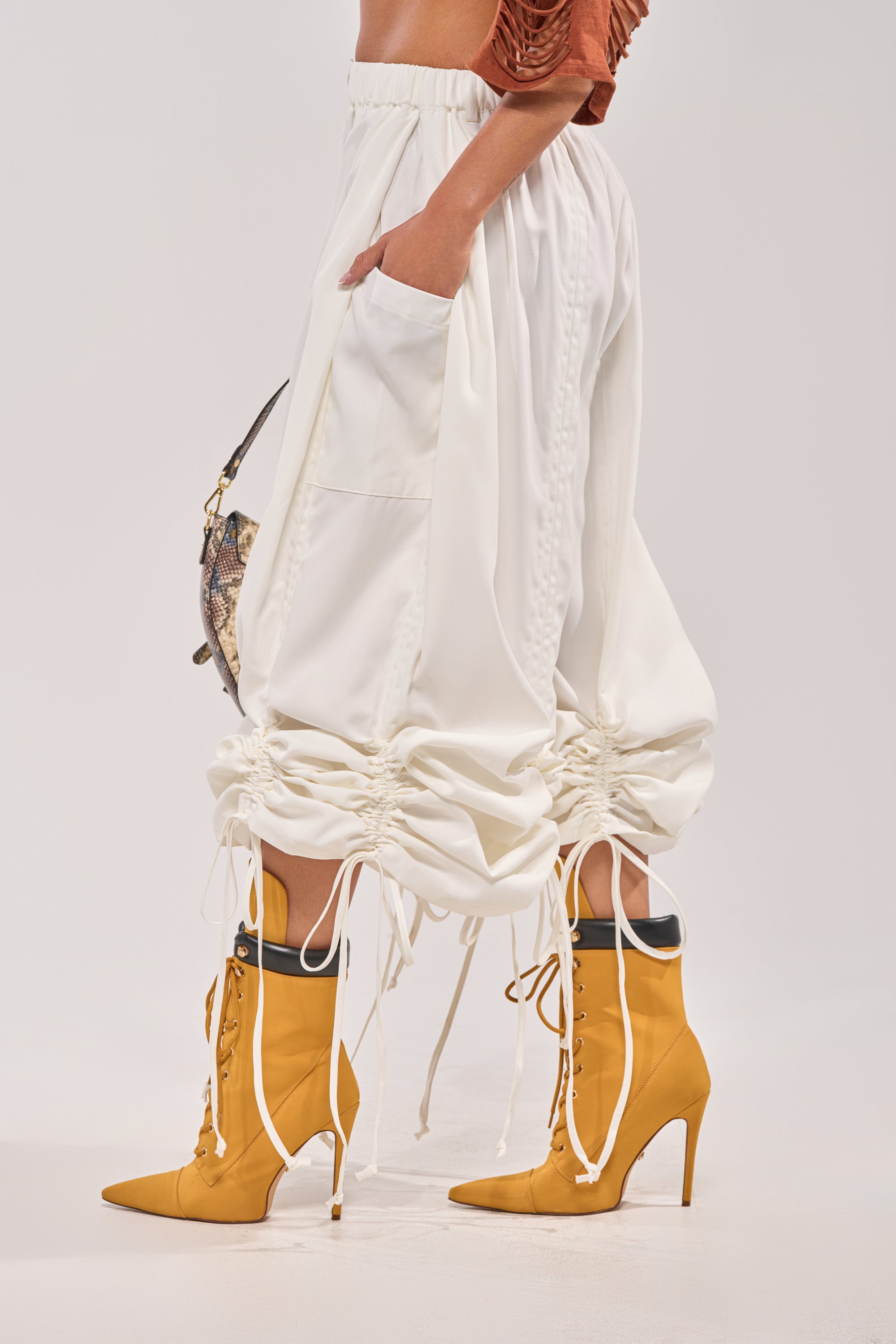 Wearing the DRAMA RUCHED JOGGER IN WHITE with gathered drawstring hems, paired with mustard yellow heeled boots, a brown cropped top, and carrying a small patterned handbag. 4