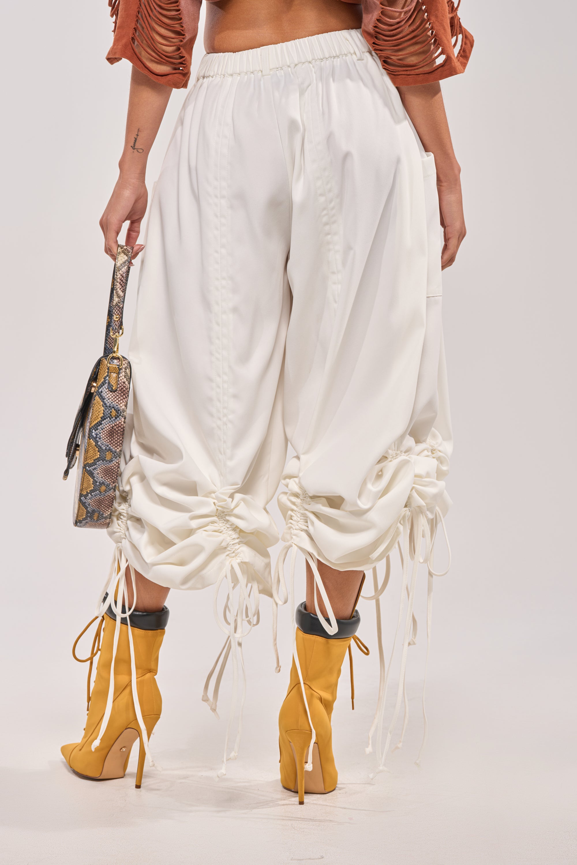A person is shown from the back, visible only from the waist down, wearing DRAMA RUCHED JOGGER IN WHITE, yellow high-heeled boots, and holding a snakeskin-patterned handbag. 5