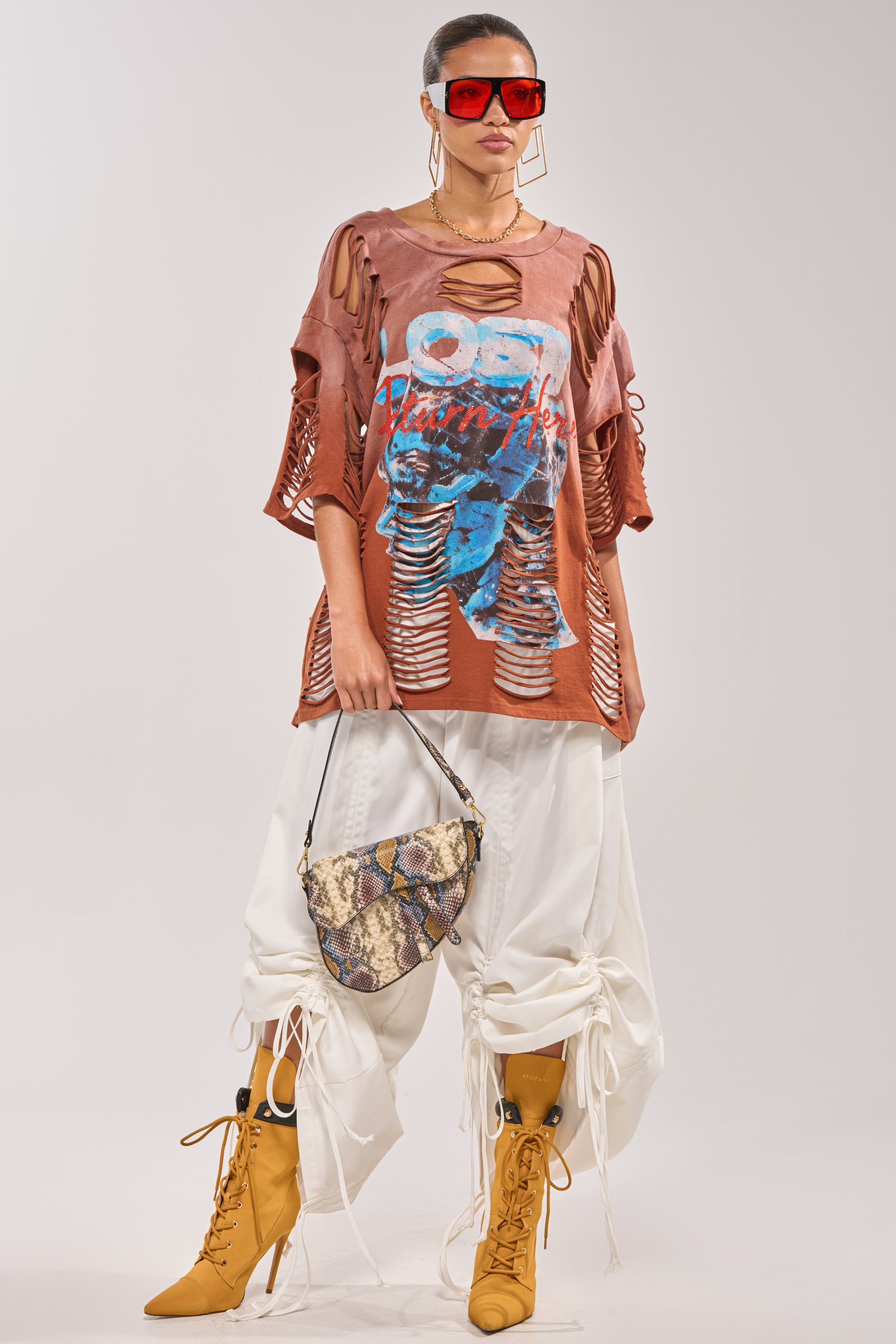 A woman wears oversized sunglasses, a distressed graphic top, the DRAMA RUCHED JOGGER IN WHITE, mustard lace-up boots, and carries a snakeskin handbag. Her hair is pulled back and she finishes the look with large hoop earrings. 6