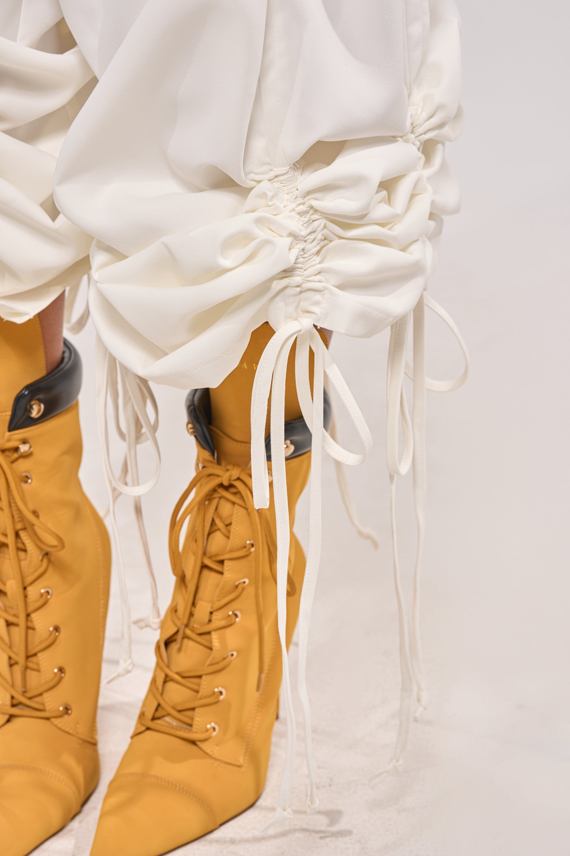 Close-up of a person wearing the DRAMA RUCHED JOGGER IN WHITE with drawstring cuffs and tan lace-up heeled boots, showcasing effortless street style against a neutral, minimal background. 7