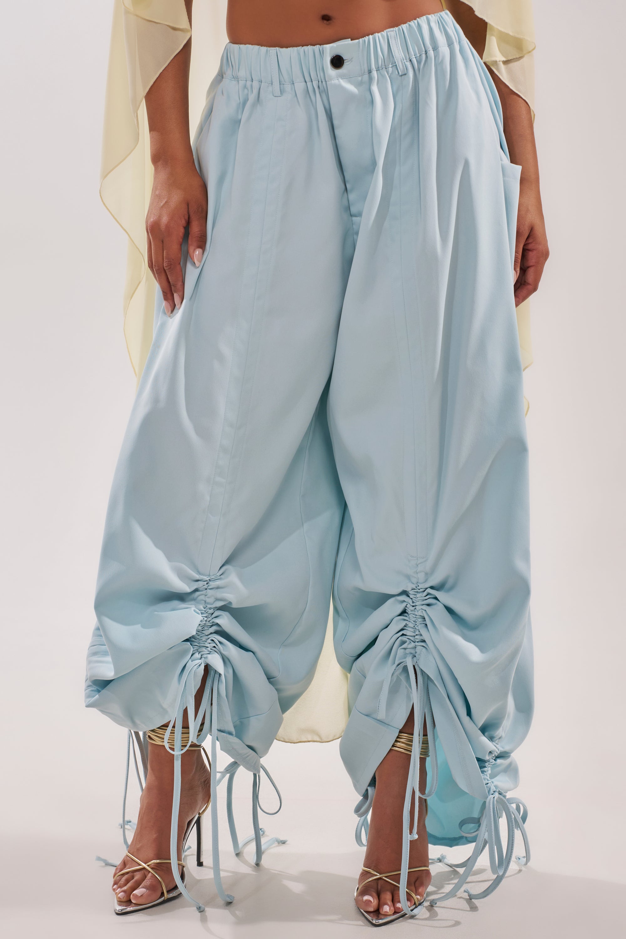 A person wearing the DRAMA RUCHED JOGGER IN BLUE with adjustable ankle drawstrings, styled with sheer high-heeled sandals and a flowing beige top—a chic, modern outfit ideal for resort vibes. 0
