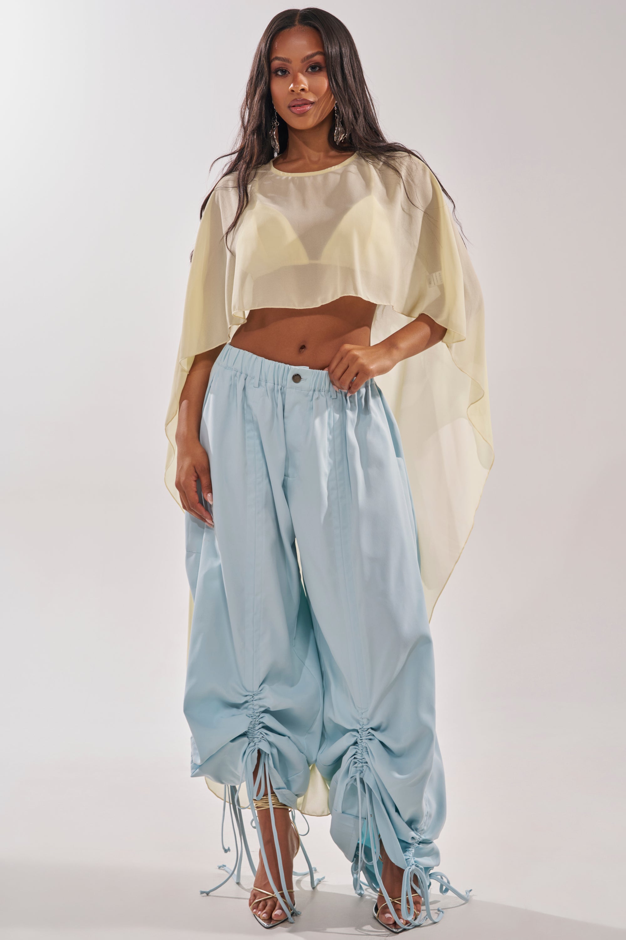 A woman models the DRAMA RUCHED JOGGER IN BLUE, high-waisted with adjustable ruching, paired with a sheer cream crop top and heels, standing against a white background. 9