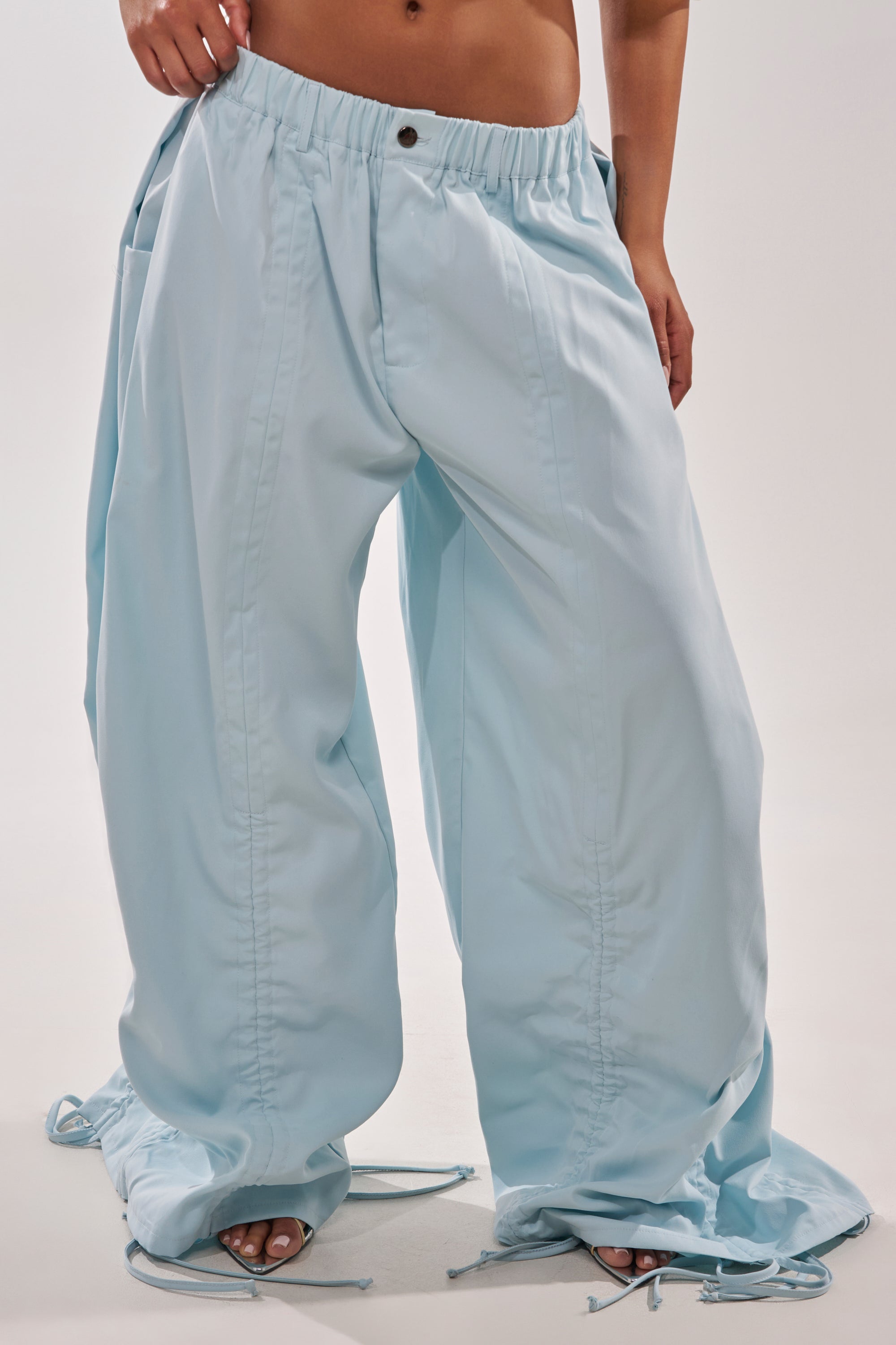 A person stands barefoot against a plain background, wearing the DRAMA RUCHED JOGGER IN BLUE—light blue, high-waisted joggers featuring adjustable drawstring ruching at the hem. 11