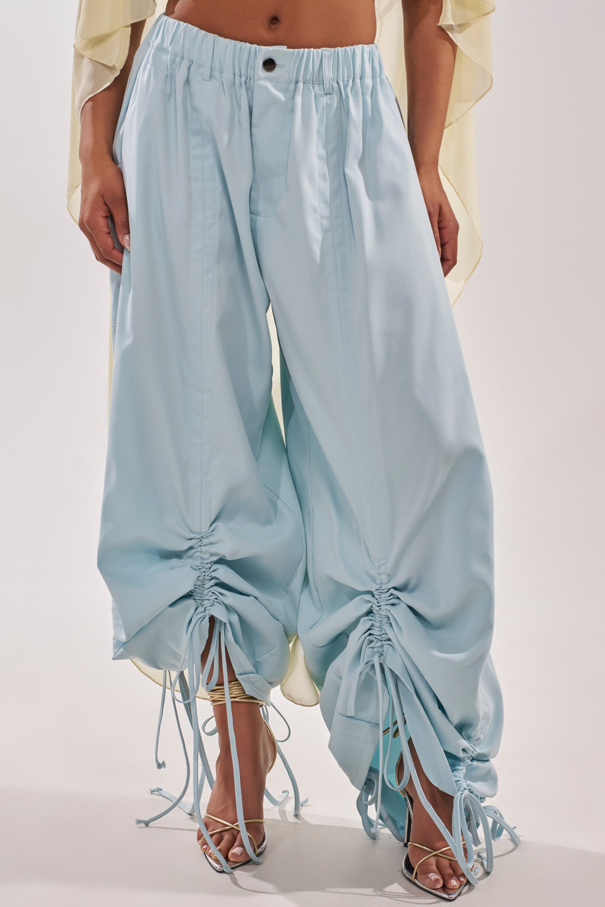 A person wears the DRAMA RUCHED JOGGER IN BLUE—loose, high-waisted wide-leg pants with ankle drawstrings—paired with silver high-heeled sandals against a plain light background for an effortless resort look. 1