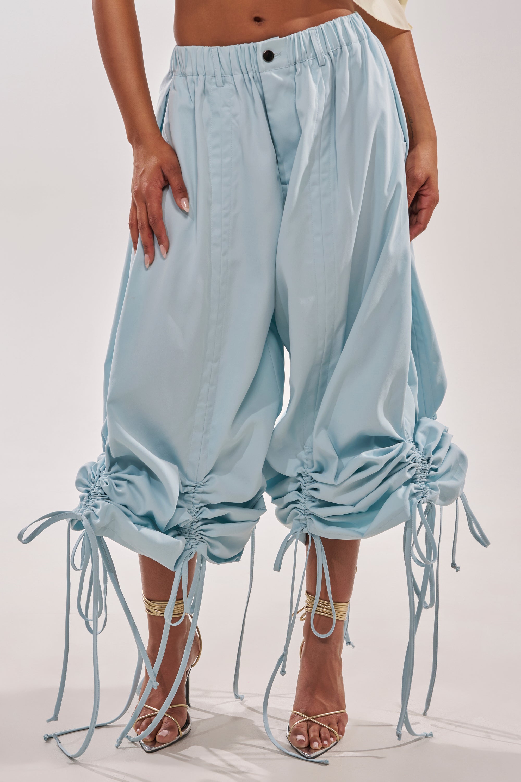 Showcasing resort style, a person wears the DRAMA RUCHED JOGGER IN BLUE with strappy nude high-heeled sandals, standing against a plain white background. 2