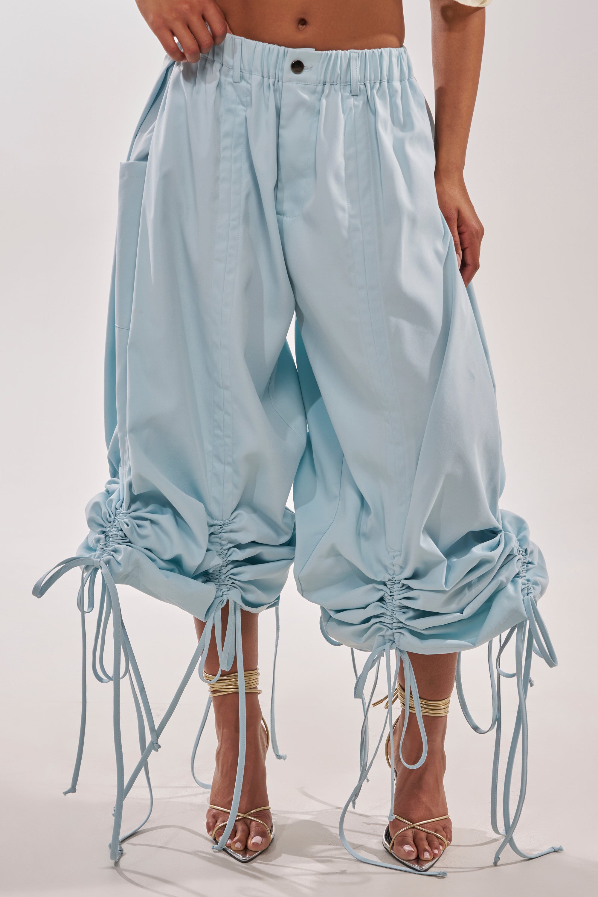 Wearing the DRAMA RUCHED JOGGER IN BLUE with drawstrings at the knees and ankles, paired with strappy heels, this look channels effortless resort style. The person's upper body is partially visible. 3
