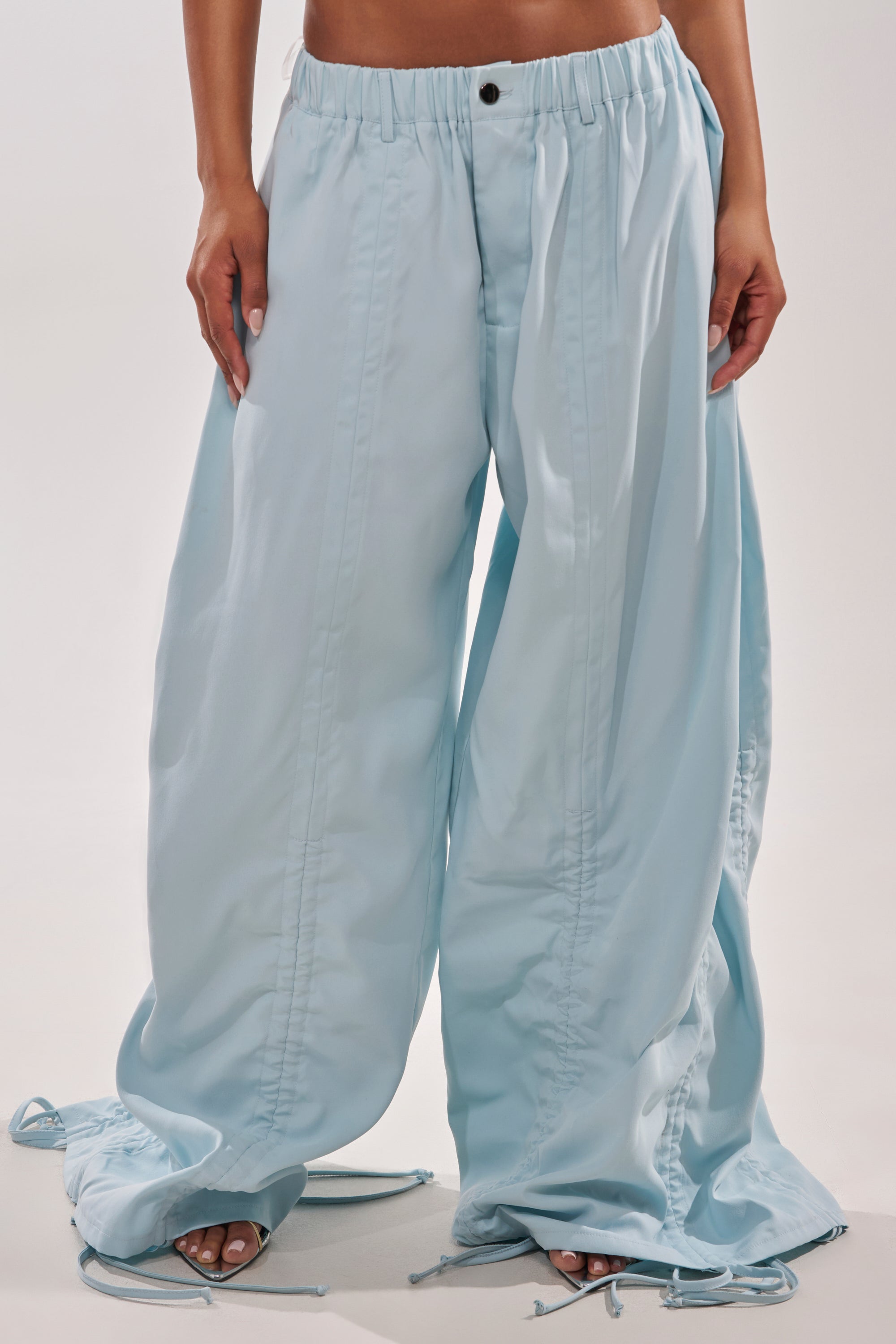 Wearing the DRAMA RUCHED JOGGER IN BLUE, featuring an elastic waistband and relaxed, oversized wide-leg fit that covers the feet. The light blue high-waisted trousers are shown from lower torso to feet against a plain background. 4