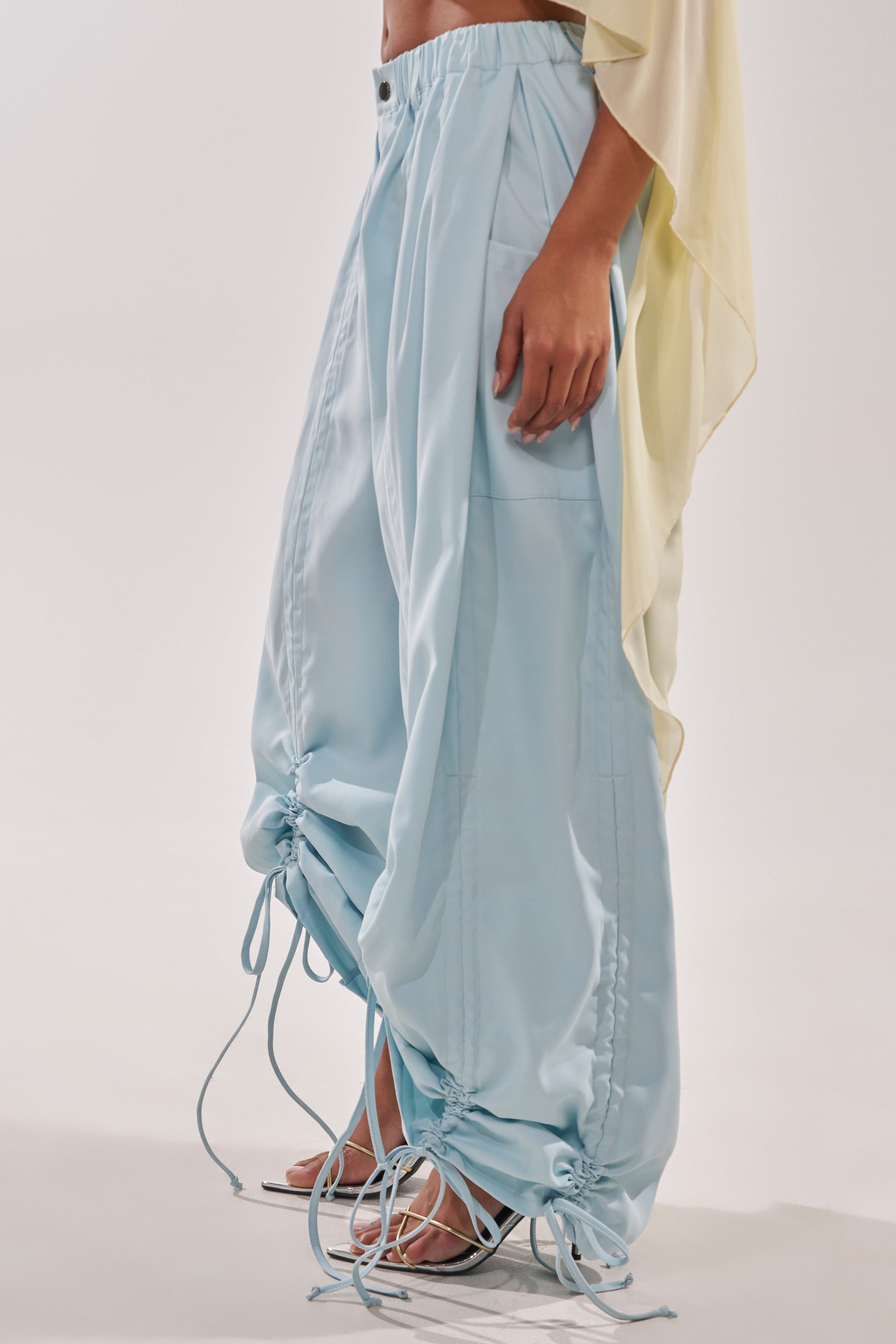Someone stands against a light background wearing the DRAMA RUCHED JOGGER IN BLUE, featuring adjustable drawstring ruching at the hem, paired with a flowy pale yellow top and white open-toe heels. 5