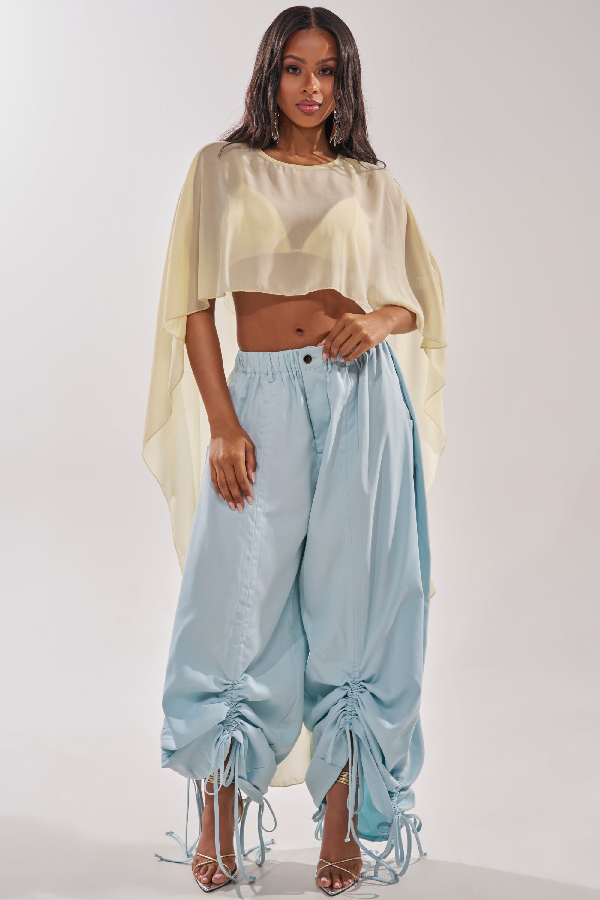 A woman wears the DRAMA RUCHED JOGGER IN BLUE, featuring adjustable drawstring ruching, paired with a sheer yellow cropped top with cape sleeves and strappy heels. 7