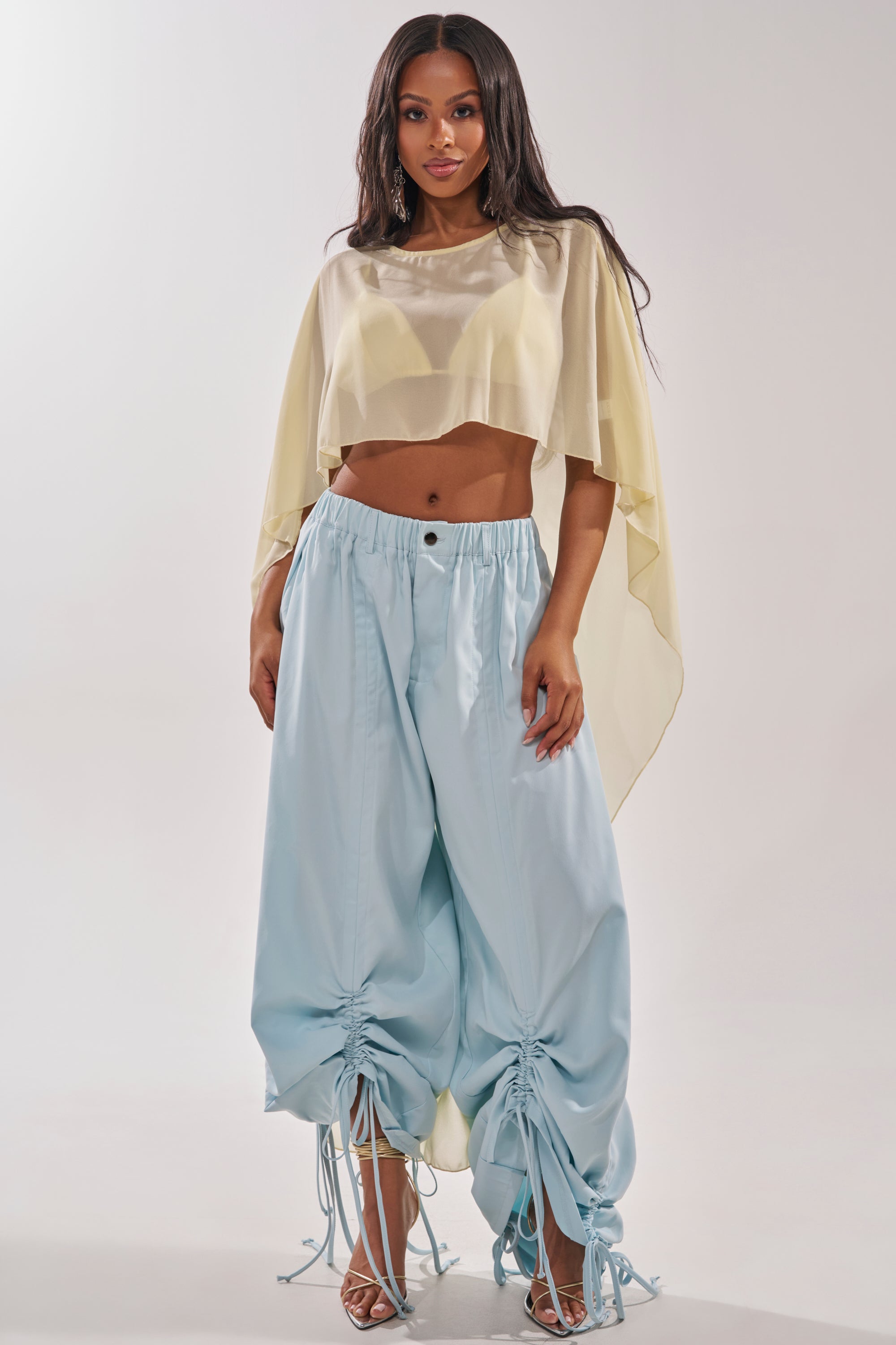 A woman poses in a sheer, pale yellow cropped cape top and the DRAMA RUCHED JOGGER IN BLUE, styled with open-toe heels against a plain, light background. 8
