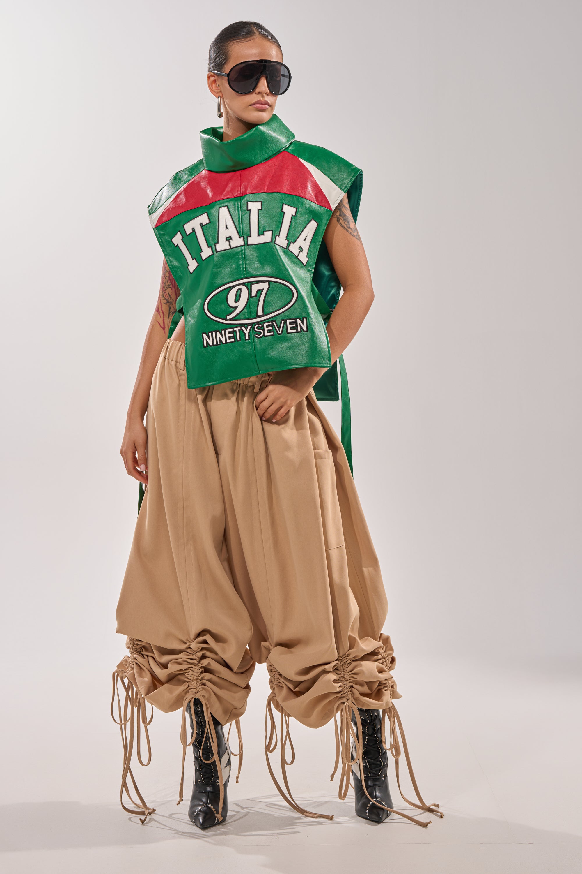 A person poses confidently against a plain background, wearing oversized black sunglasses, a green and red sleeveless "ITALIA 97 NINETY SEVEN" top, black boots, and the DRAMA RUCHED JOGGER IN KHAKI. 1