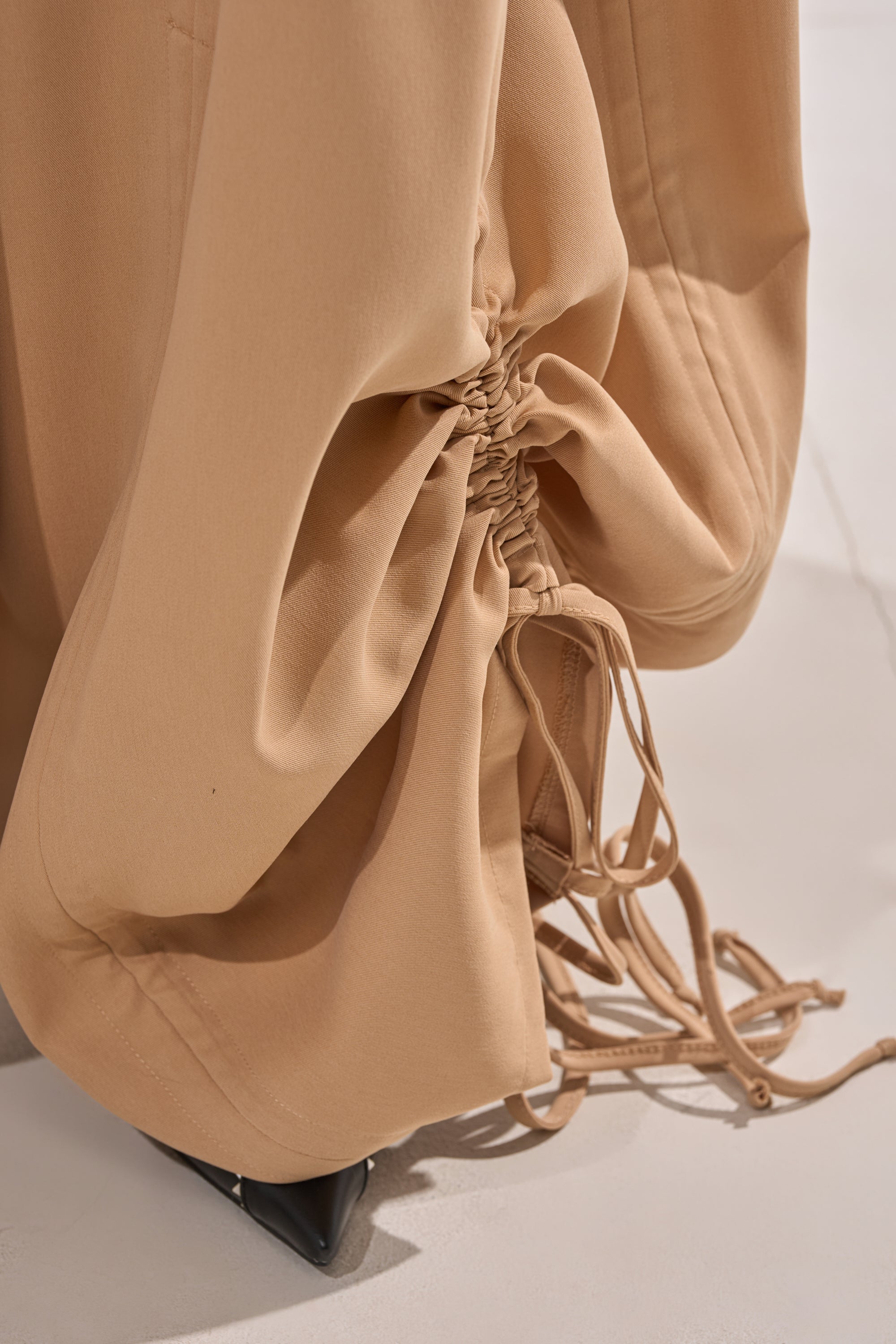 The DRAMA RUCHED JOGGER IN KHAKI features gathered ruching and cinching drawstrings, stylishly draped over pointed black shoes on a light floor. 10