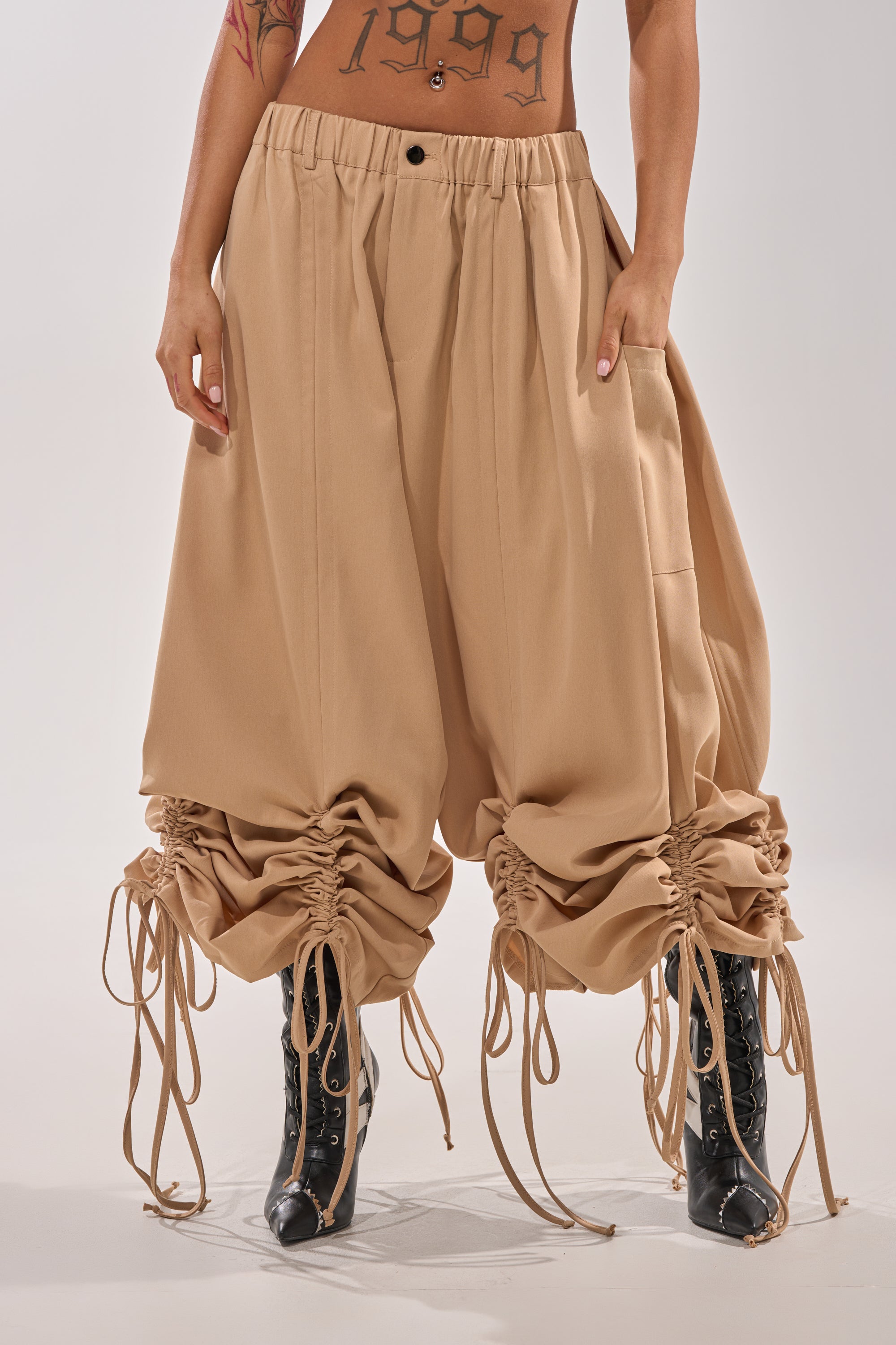 Someone wears the DRAMA RUCHED JOGGER IN KHAKI with drawstring ankles, paired with black lace-up boots. The high-rise, oversized joggers showcase a streetwear vibe, and tattoos including "1999" are visible on their torso. 2