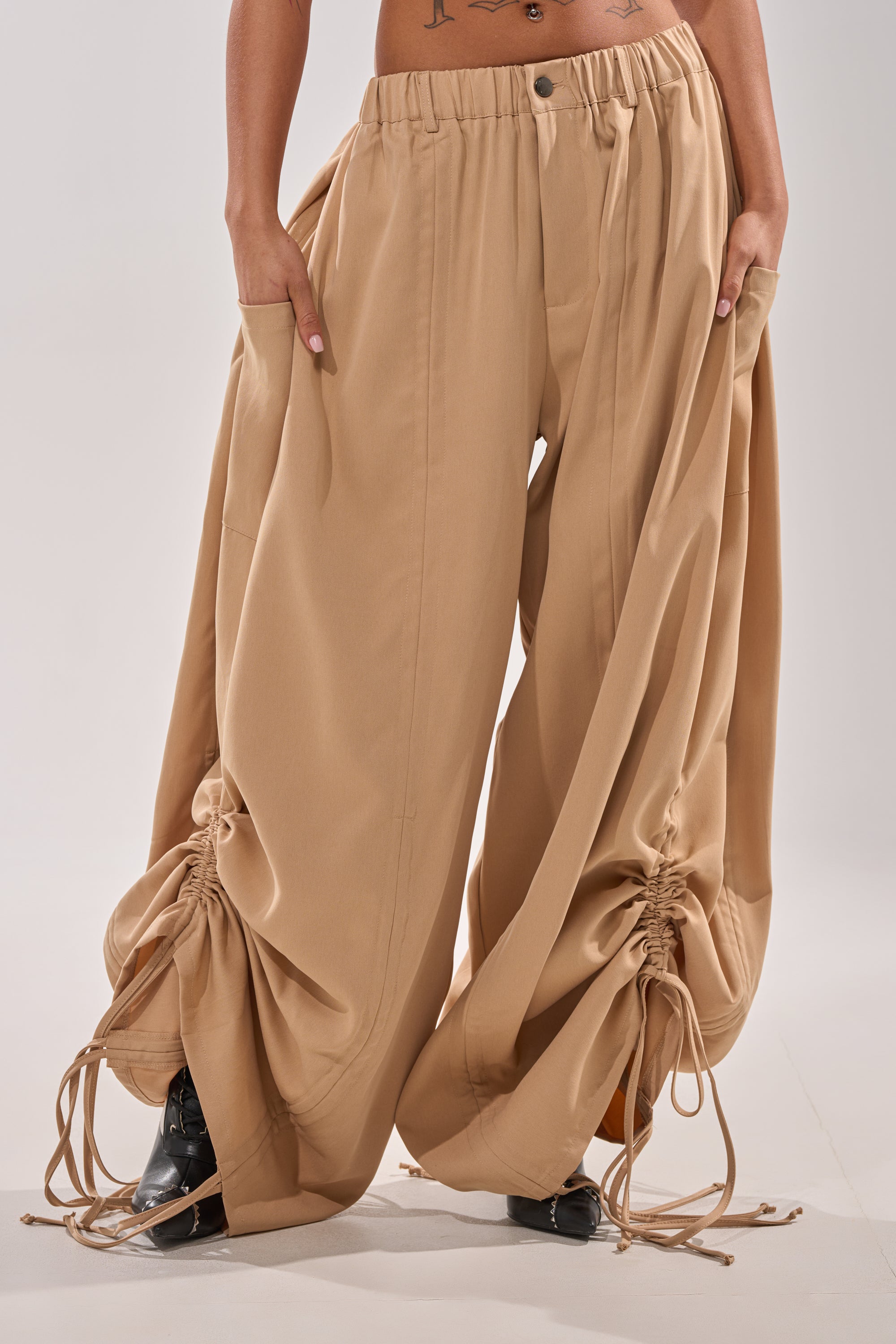 Model wears the DRAMA RUCHED JOGGER IN KHAKI, featuring a high-rise elastic waistband and gathered ankles. Only the lower torso and legs are shown, hands in pockets, with tattoos visible on the lower abdomen. Paired with black shoes. 0
