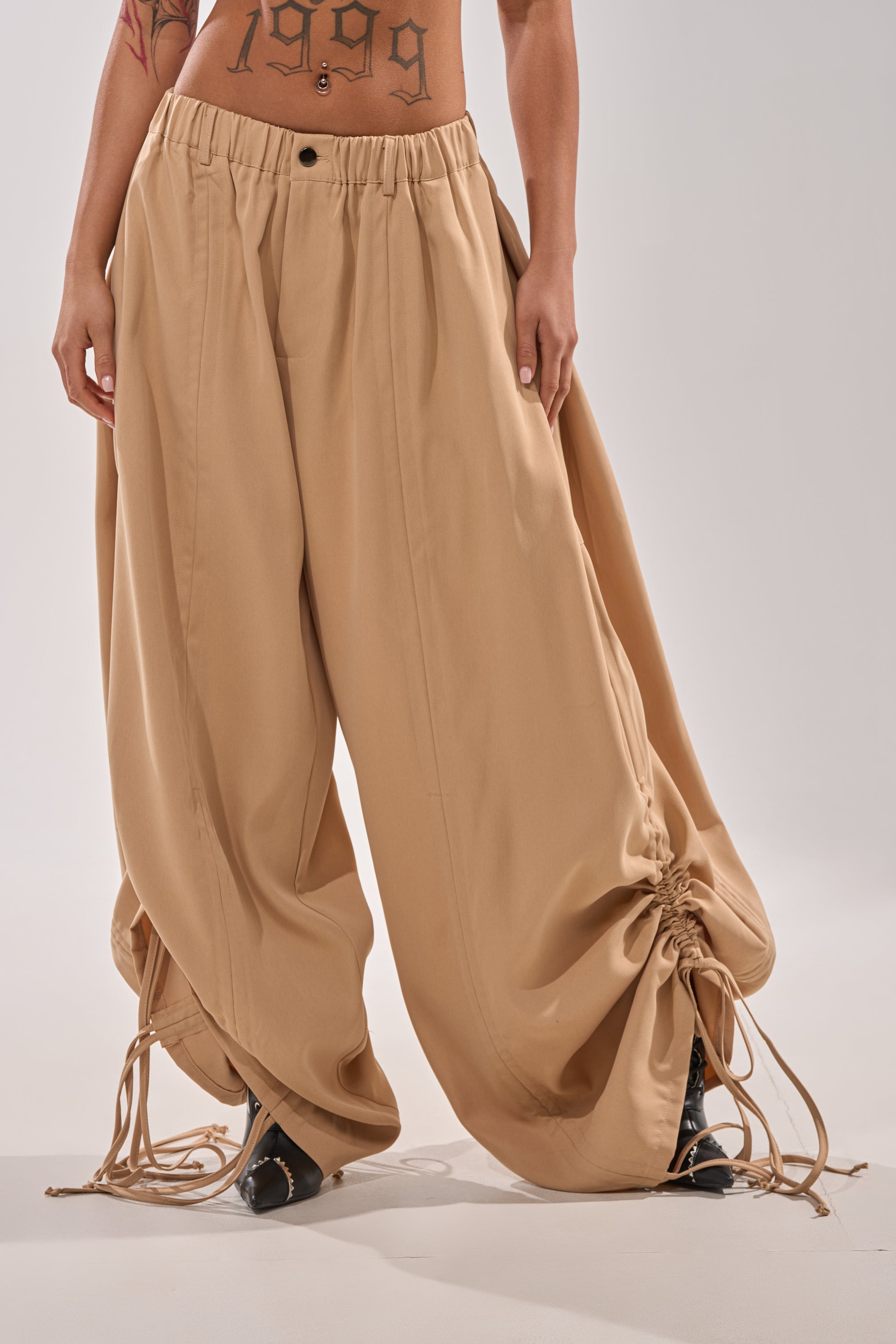 A person wears the DRAMA RUCHED JOGGER IN KHAKI with drawstring and ruched ankles, revealing a “1999” stomach tattoo. Black pointed shoes finish this bold street style look. 3